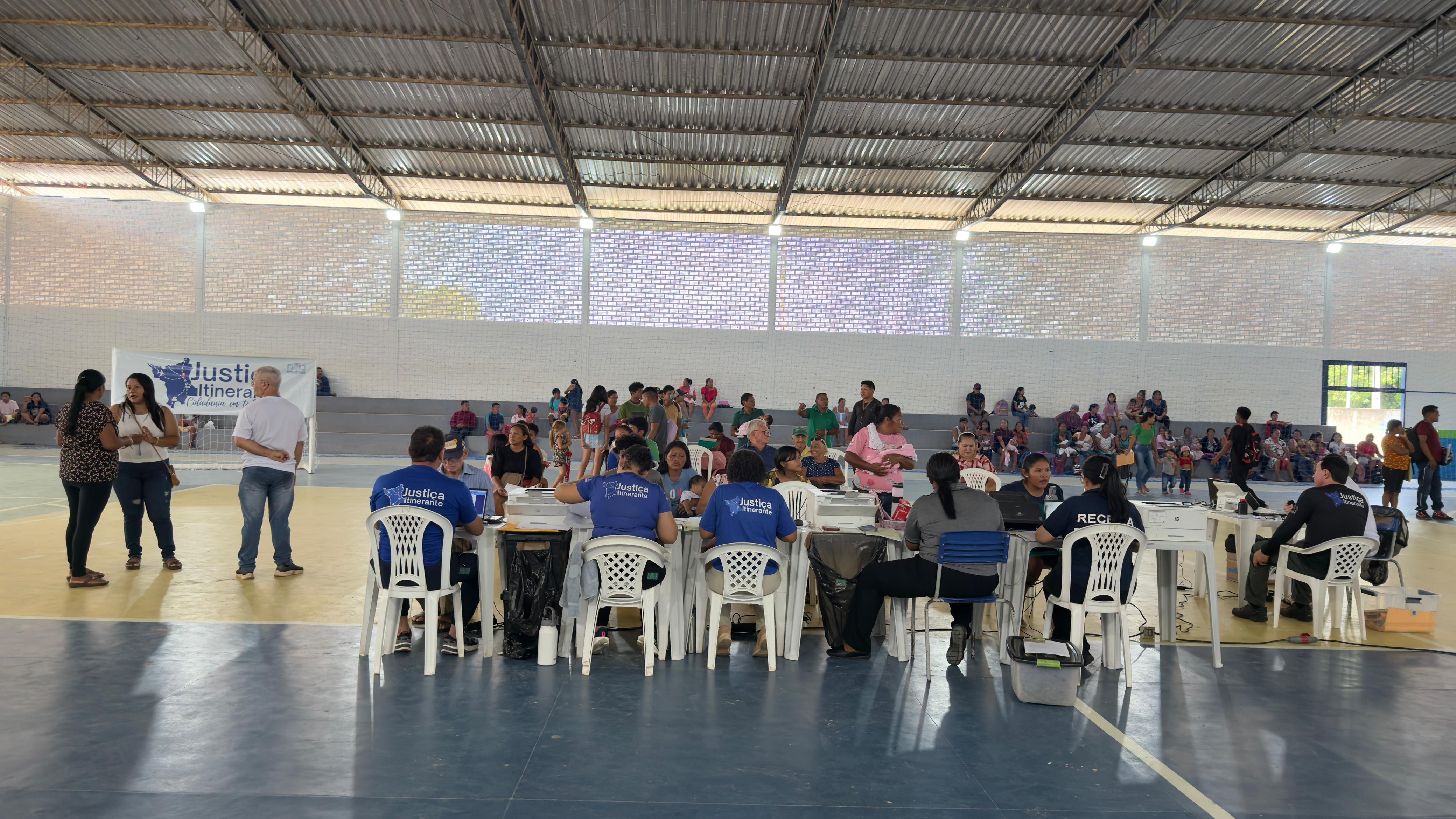 A imagem colorida mostra um ginásio ou quadra coberta onde um evento está ocorrendo. Há uma mesa com várias pessoas sentadas de costas para a câmera, trabalhando com computadores e documentos, possivelmente prestando algum tipo de serviço. No fundo e nas laterais, há um grande número de pessoas em pé e sentadas, algumas em grupos, outras aguardando. Uma faixa ou banner com a palavra "Justiça" é visível no lado esquerdo. A imagem colorida mostra um ginásio ou quadra coberta onde um evento está ocorrendo. Há uma mesa com várias pessoas sentadas de costas para a câmera, trabalhando com computadores e documentos, possivelmente prestando algum tipo de serviço. No fundo e nas laterais, há um grande número de pessoas em pé e sentadas, algumas em grupos, outras aguardando. Uma faixa ou banner com a palavra "Justiça" é visível no lado esquerdo.