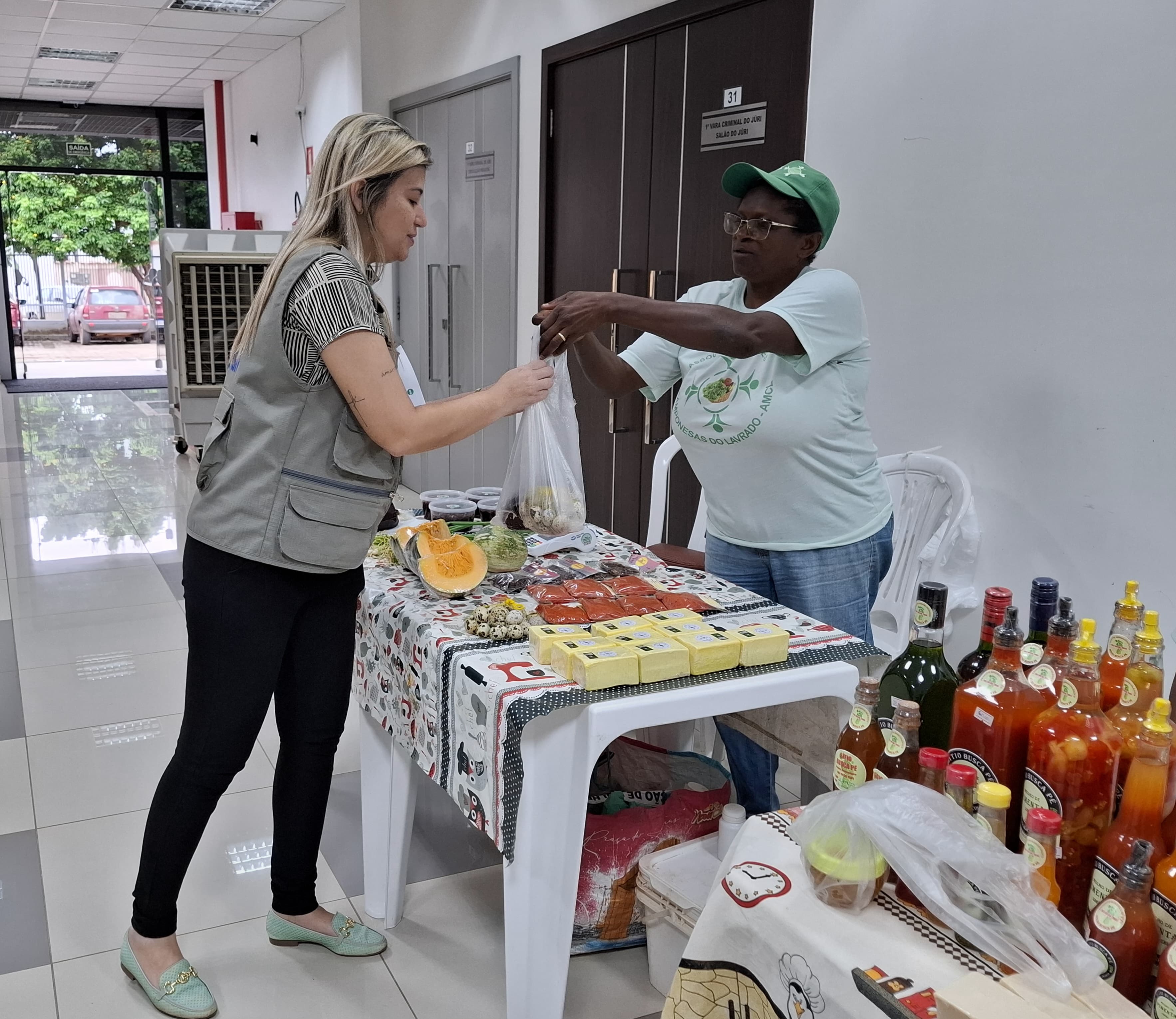 A imagem colorida mostra uma mulher entregando uma sacola com produtos para uma outra mulher de colete. Ambas estão ao redor de uma mesa, sobre a qual há diversos produtos, incluindo queijos embalados e garrafas de bebidas. A imagem colorida mostra uma mulher entregando uma sacola com produtos para uma outra mulher de colete. Ambas estão ao redor de uma mesa, sobre a qual há diversos produtos, incluindo queijos embalados e garrafas de bebidas.