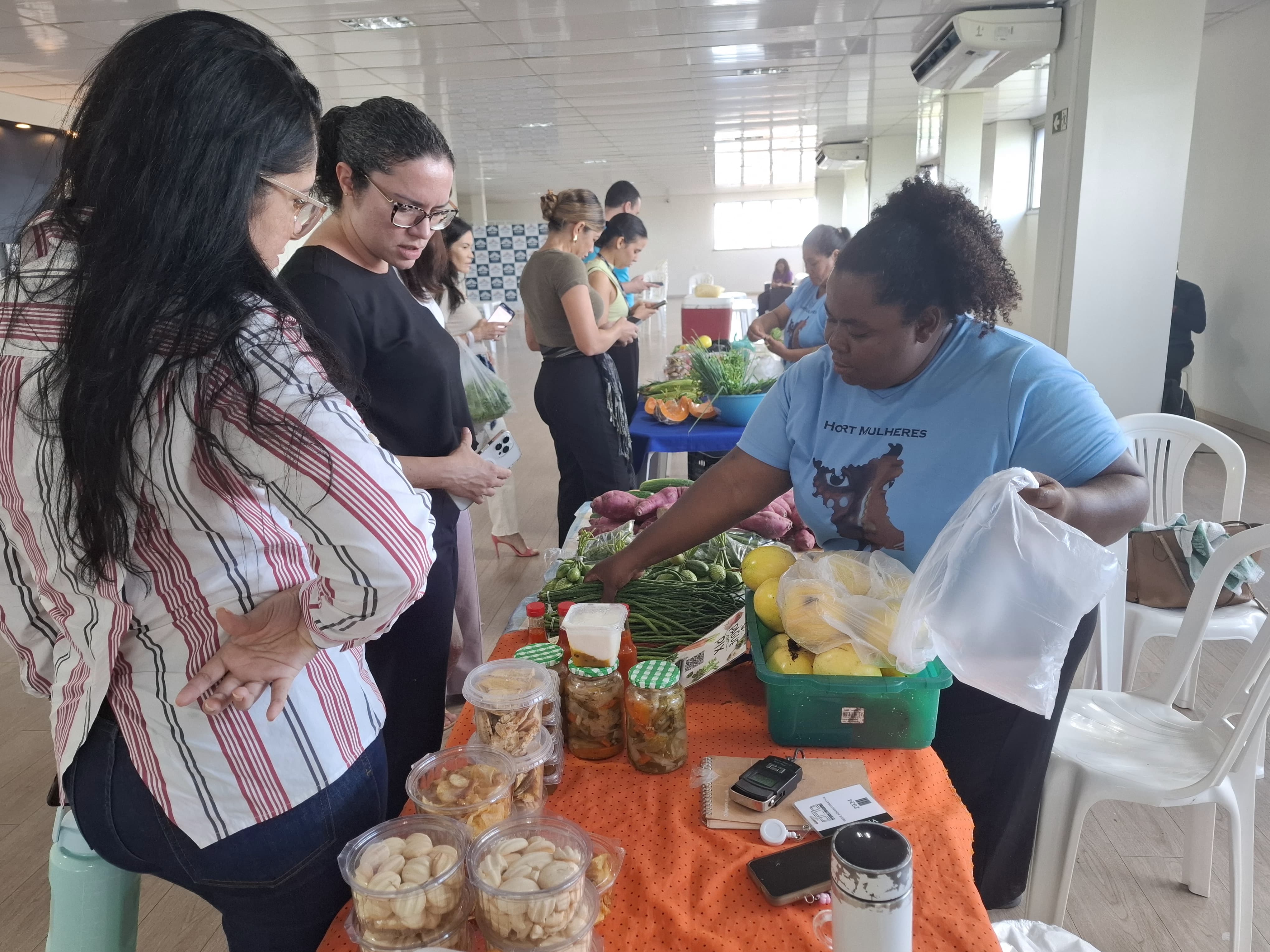 A imagem colorida mostra um ambiente interno onde uma mulher com uma camiseta azul atende duas mulheres. Há uma mesa com produtos como vegetais frescos, potes de doces ou conservas e outros itens embalados. Ao fundo, outras mulheres também estão sendo atendidas. A imagem colorida mostra um ambiente interno onde uma mulher com uma camiseta azul atende duas mulheres. Há uma mesa com produtos como vegetais frescos, potes de doces ou conservas e outros itens embalados. Ao fundo, outras mulheres também estão sendo atendidas.