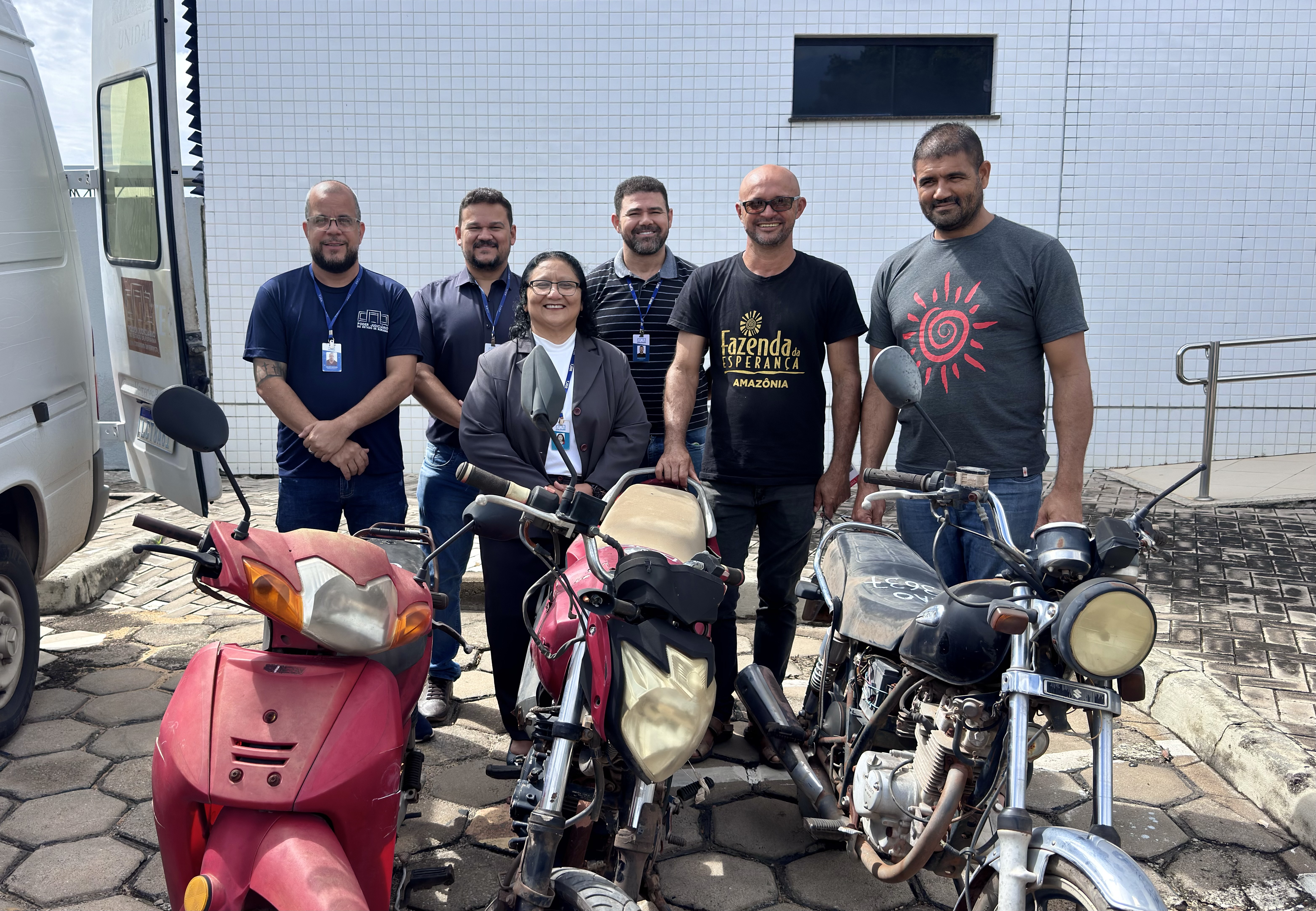 Imagem colorida mostra seis pessoas estão em pé, sorrindo, posicionadas atrás de três motocicletas antigas. À esquerda, há uma van branca. Ao fundo, uma parede de azulejos brancos com uma pequena janela escura. Duas das pessoas vestem camisetas com a inscrição “Fazenda da Esperança Amazônia”, e os demais usam crachás de identificação. A cena acontece ao ar livre, em um pátio com piso de pedras. Imagem colorida mostra seis pessoas estão em pé, sorrindo, posicionadas atrás de três motocicletas antigas. À esquerda, há uma van branca. Ao fundo, uma parede de azulejos brancos com uma pequena janela escura. Duas das pessoas vestem camisetas com a inscrição “Fazenda da Esperança Amazônia”, e os demais usam crachás de identificação. A cena acontece ao ar livre, em um pátio com piso de pedras.