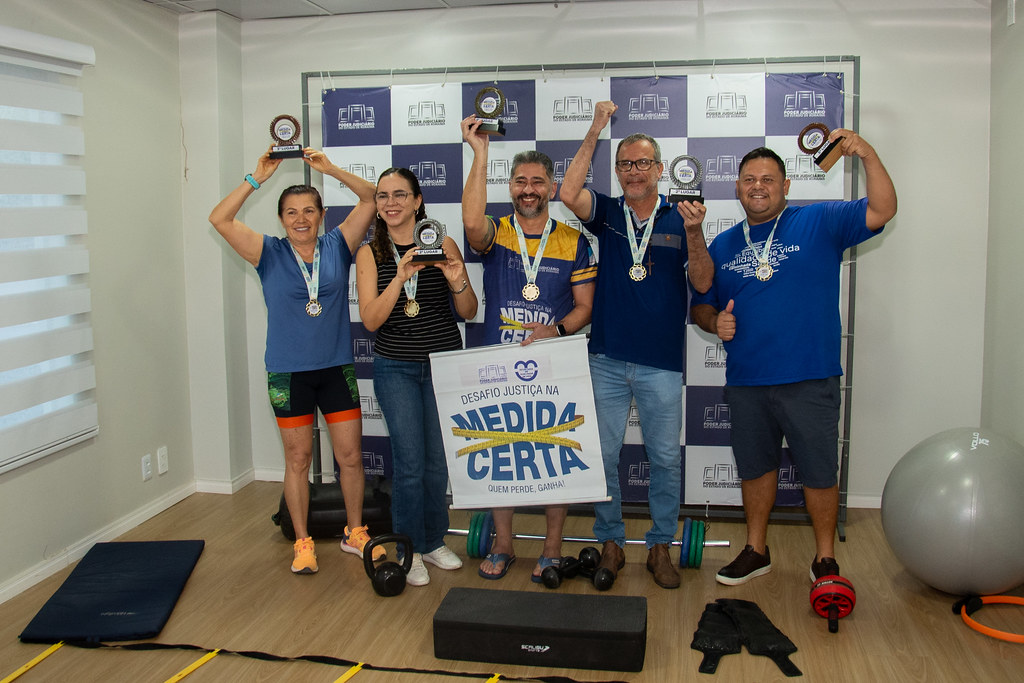 Cinco participantes do desafio posam animados com troféus e medalhas, exibindo sorrisos e gestos de comemoração. Eles seguram um banner do programa e estão em um espaço de atividade física com colchonetes, halteres e equipamentos de treino. Cinco participantes do desafio posam animados com troféus e medalhas, exibindo sorrisos e gestos de comemoração. Eles seguram um banner do programa e estão em um espaço de atividade física com colchonetes, halteres e equipamentos de treino.