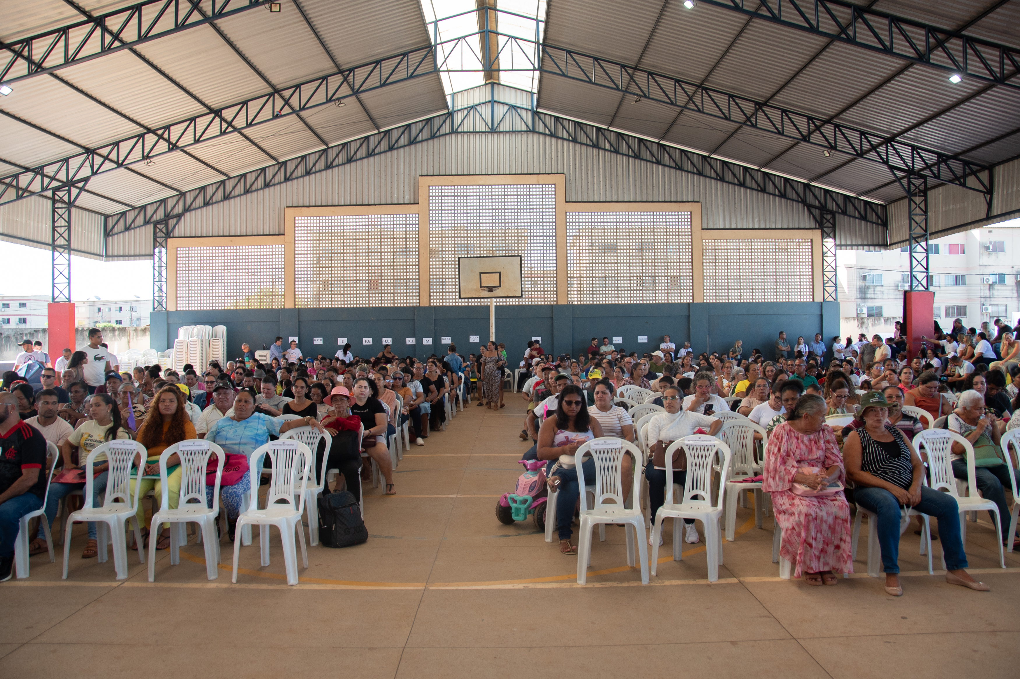 Foto colorida do ginásio da escola com pessoas sentadas