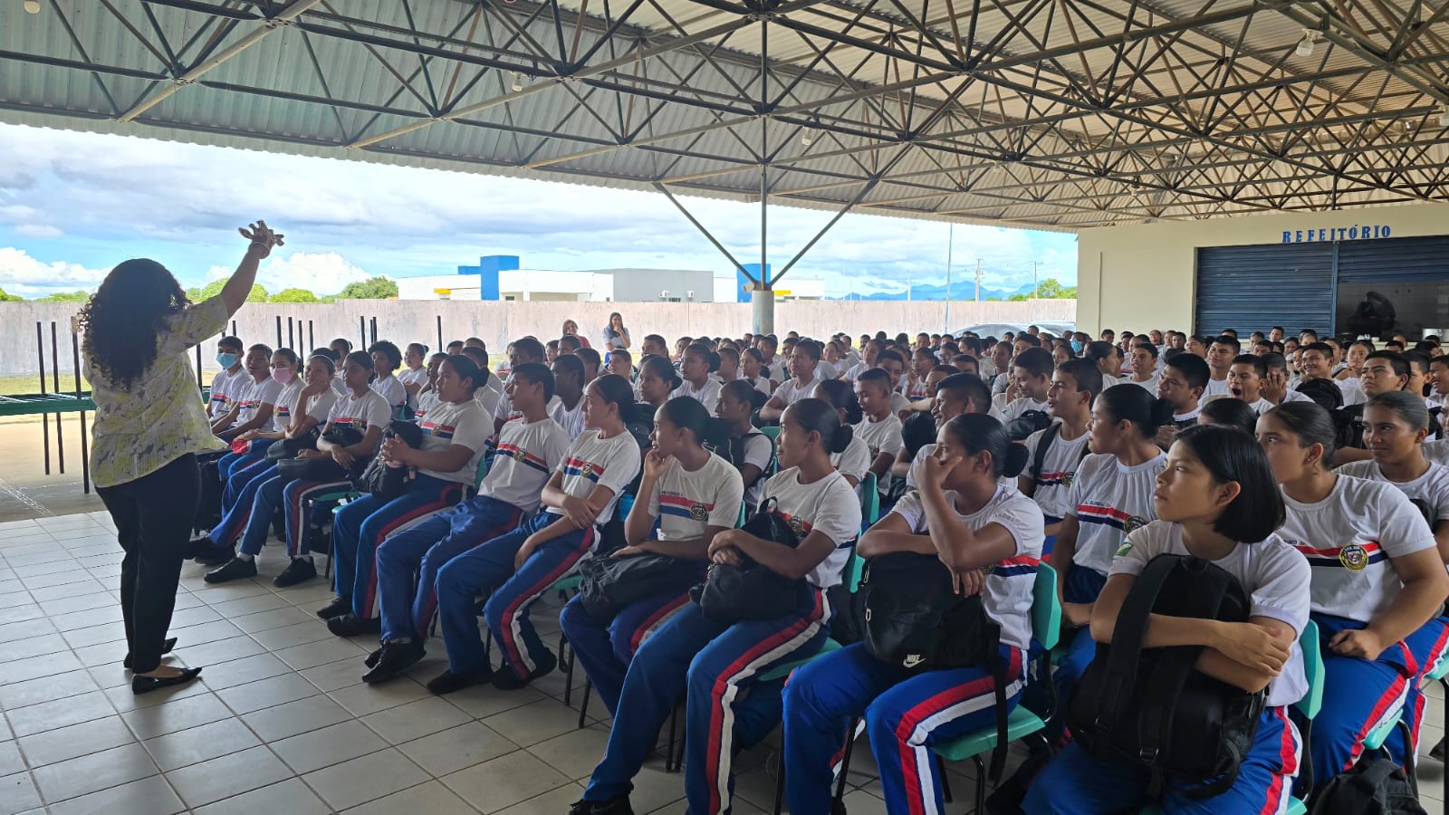 A imagem colorida mostra uma mulher de costas para a câmera, com o braço levantado, falando para uma grande plateia de jovens sentados em cadeiras. Os jovens, que parecem ser estudantes, vestem uniformes brancos com listras azuis e vermelhas e estão sentados sob uma estrutura coberta, com o céu e prédios ao fundo. Há uma placa visível ao fundo com a palavra "REFEITÓRIO". A imagem colorida mostra uma mulher de costas para a câmera, com o braço levantado, falando para uma grande plateia de jovens sentados em cadeiras. Os jovens, que parecem ser estudantes, vestem uniformes brancos com listras azuis e vermelhas e estão sentados sob uma estrutura coberta, com o céu e prédios ao fundo. Há uma placa visível ao fundo com a palavra "REFEITÓRIO".