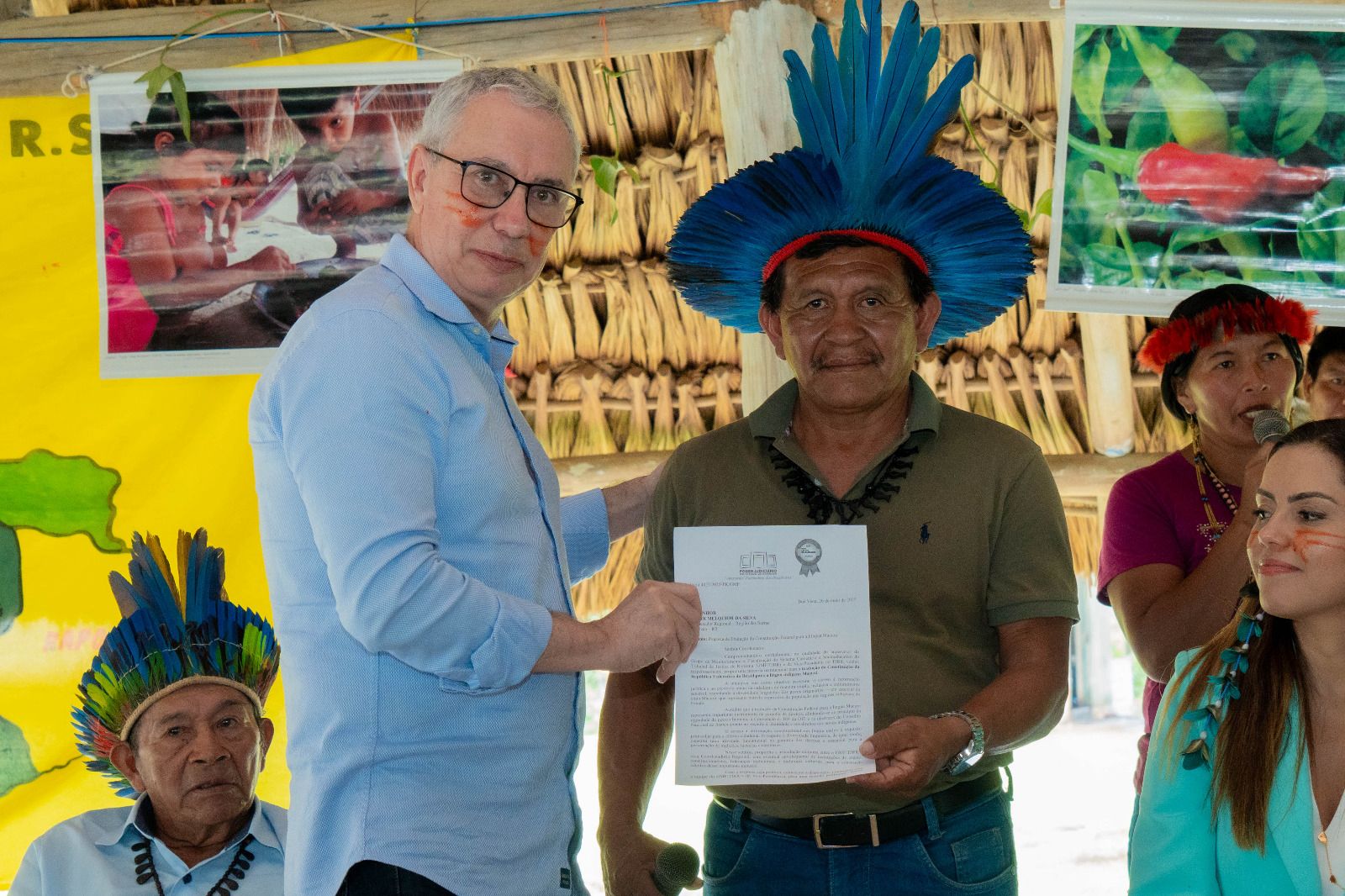 A imagem colorida mostra o Desembargador Almiro Padilha ao lado do Tuxaua regional Djacir Melquior segurando um documento, durante a ação inédita da tradução da Constituição Federal para a língua Macuxi. Atrás deles encontram-se um banner e uma mulher da comunidade falando no microfone. 