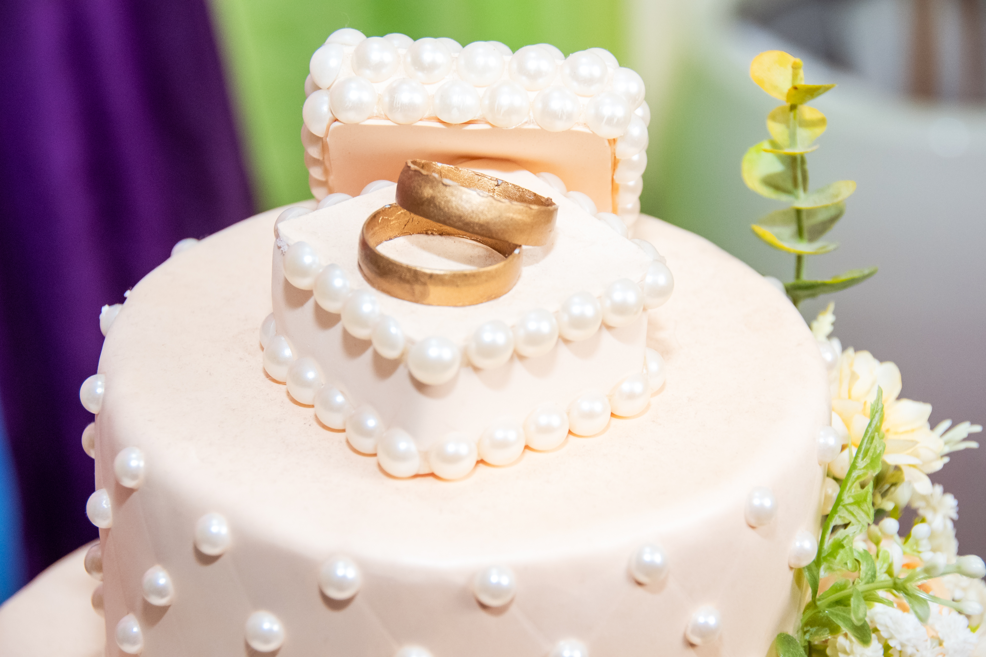 A imagem colorida mostra uma foto de perto de um bolo de casamento branco com detalhes que imitam pérolas. No topo do bolo, há duas alianças douradas. A imagem colorida mostra uma foto de perto de um bolo de casamento branco com detalhes que imitam pérolas. No topo do bolo, há duas alianças douradas.