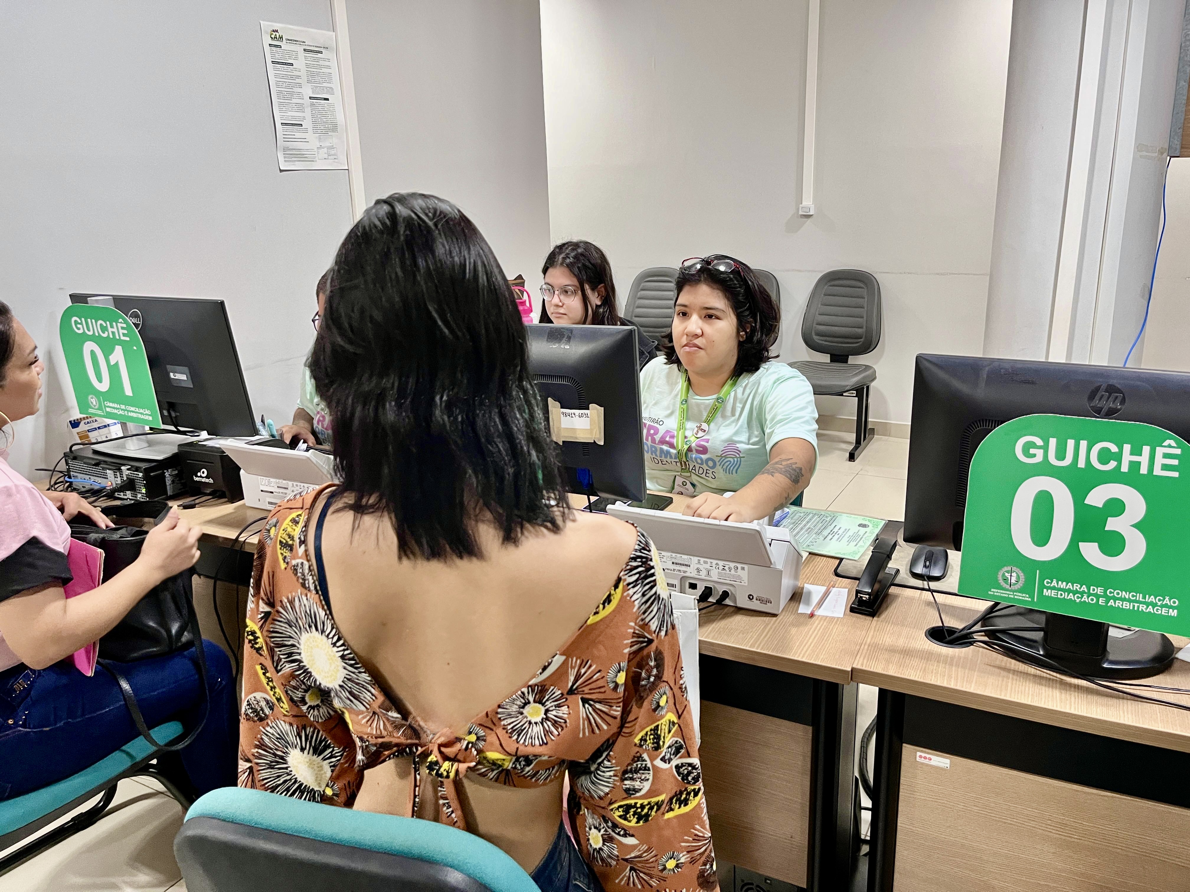 A imagem colorida mostra uma mulher de costas, sentada em uma cadeira azul, olha para três atendentes em guichês em um ambiente de escritório. O guichê 03 tem um monitor HP preto e um cartaz que diz "Câmara de Conciliação, Mediação e Arbitragem" A imagem colorida mostra uma mulher de costas, sentada em uma cadeira azul, olha para três atendentes em guichês em um ambiente de escritório. O guichê 03 tem um monitor HP preto e um cartaz que diz "Câmara de Conciliação, Mediação e Arbitragem"