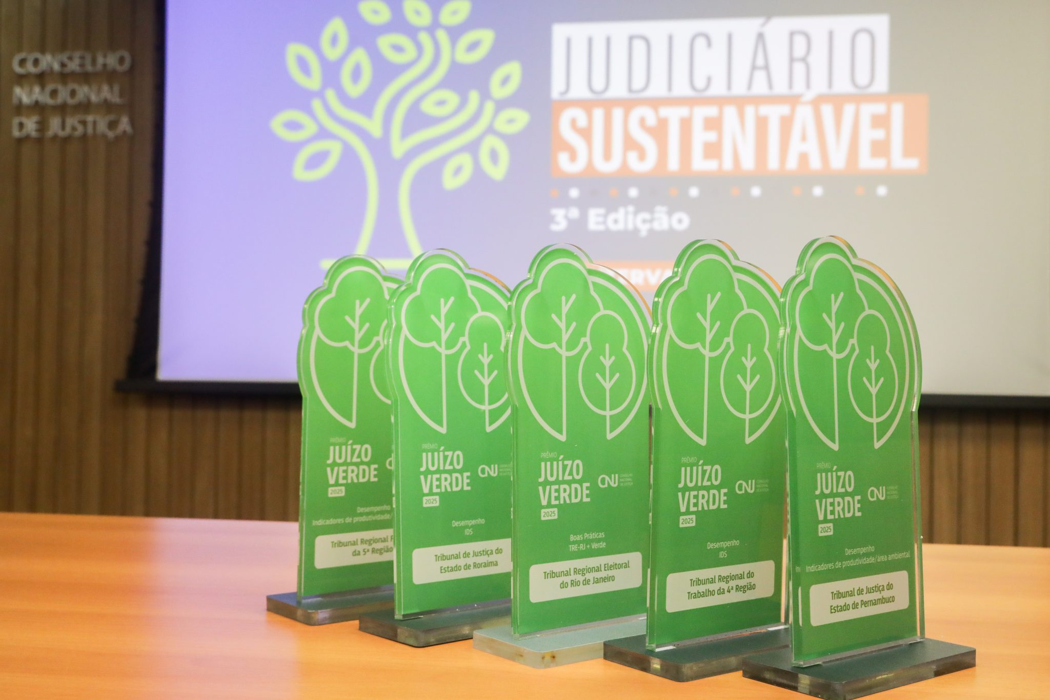 A imagem mostra uma mesa com cinco troféus verdes do "Prêmio Juízo Verde 2023", entregues a diferentes tribunais brasileiros por desempenho e boas práticas sustentáveis. Os troféus têm formato de árvore estilizada e contêm placas com inscrições específicas de cada premiação. Ao fundo, há uma tela com os dizeres "JUDICIÁRIO SUSTENTÁVEL – 3ª Edição" e o logotipo do Conselho Nacional de Justiça (CNJ), indicando que a cerimônia celebra ações sustentáveis no Judiciário.