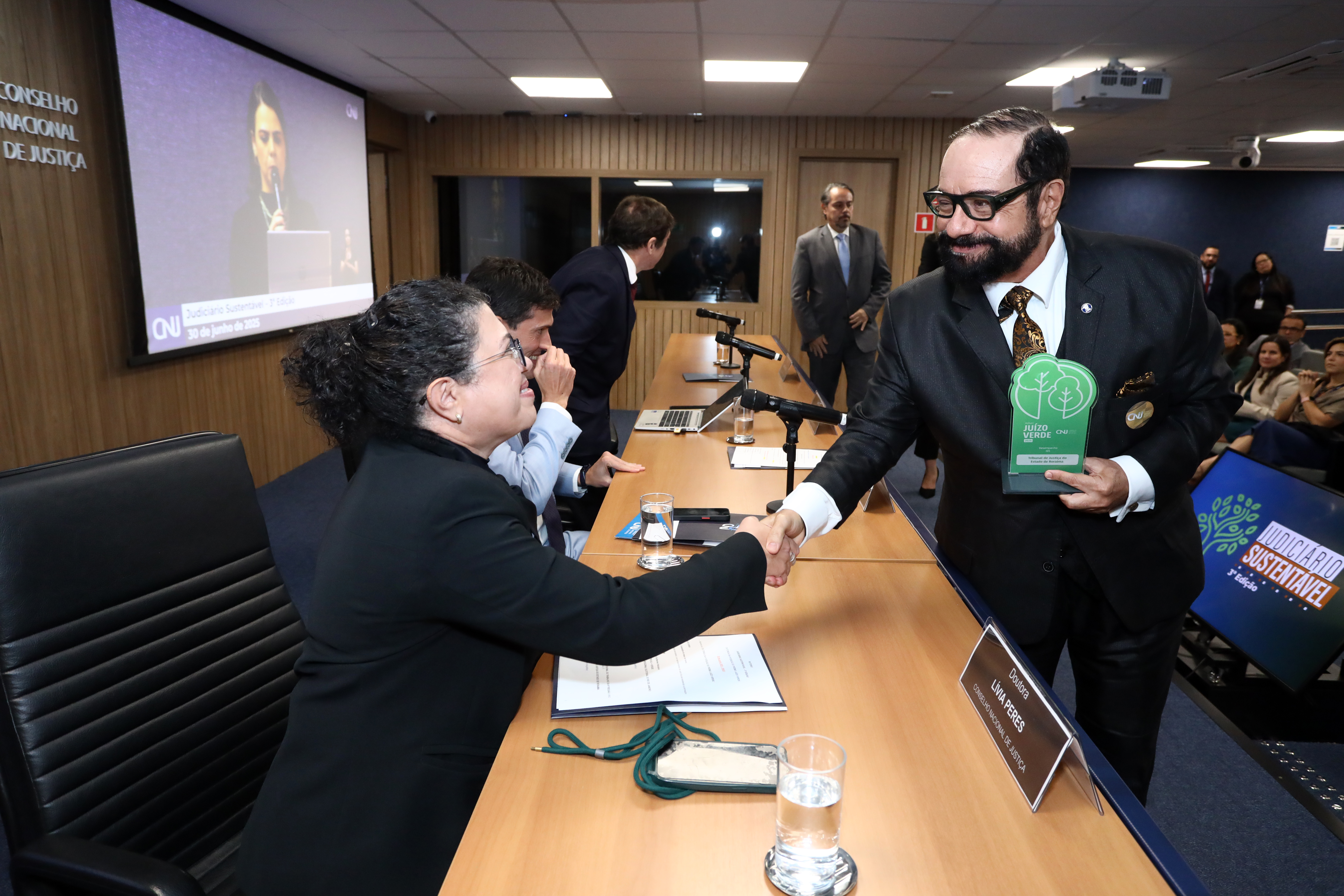 imagem colorida do presidente do TJRR, desembargador Leonardo Cupello, segurando o prêmio com a mão direita e com a mão esquerda cumprimentando uma mulher da mesa de autoridades. imagem colorida do presidente do TJRR, desembargador Leonardo Cupello, segurando o prêmio com a mão direita e com a mão esquerda cumprimentando uma mulher da mesa de autoridades.