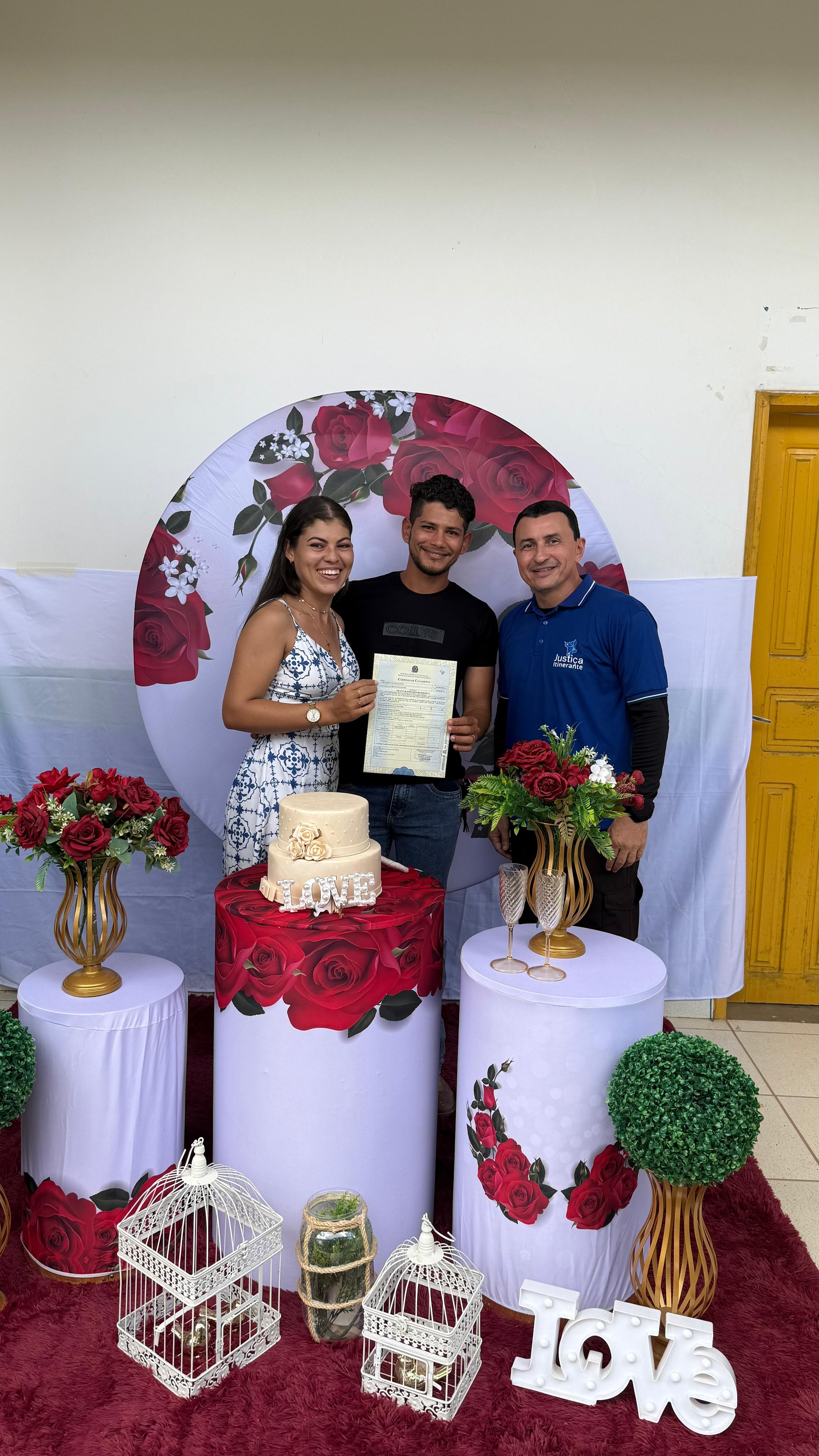 A imagem mostra um casal celebrando sua união civil durante atendimento da Vara da Justiça Itinernate. A mulher veste um vestido azul e branco, e o homem segura uma certidão de casamento, sorrindo. Ao lado deles, um servidor com camisa azul posa com a mão no ombro do noivo. Ao fundo, há uma decoração com rosas vermelhas, bolo, flores e a palavra “Love”. A cena transmite alegria e acolhimento em um ambiente festivo e simples. A imagem mostra um casal celebrando sua união civil durante atendimento da Vara da Justiça Itinernate. A mulher veste um vestido azul e branco, e o homem segura uma certidão de casamento, sorrindo. Ao lado deles, um servidor com camisa azul posa com a mão no ombro do noivo. Ao fundo, há uma decoração com rosas vermelhas, bolo, flores e a palavra “Love”. A cena transmite alegria e acolhimento em um ambiente festivo e simples.