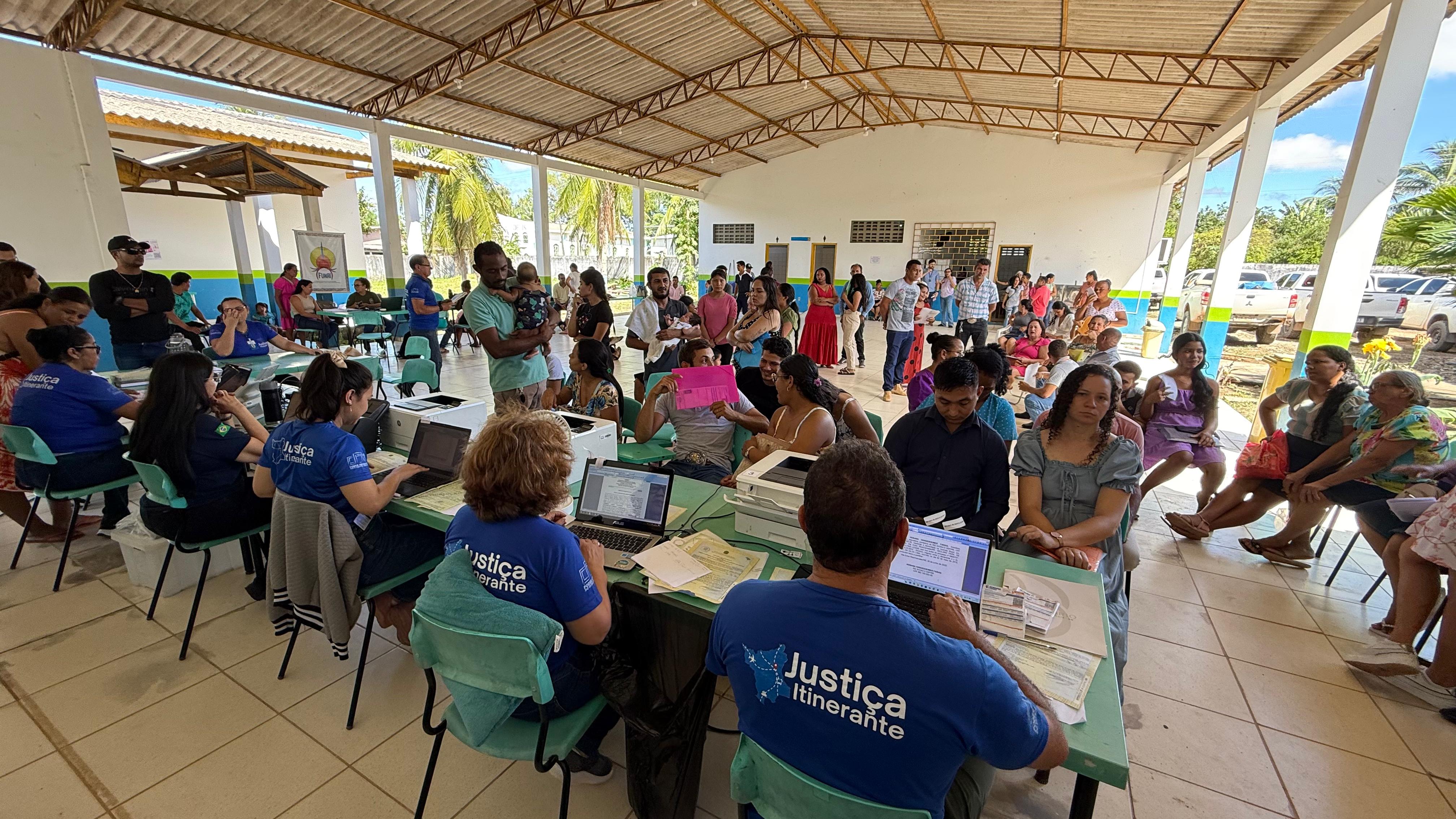 A imagem mostra um evento da Justiça Itinerante acontecendo durante o dia em um espaço aberto e coberto, com palmeiras ao fundo. Em destaque, duas pessoas de camiseta azul com a inscrição "Justiça Itinerante" atendem cidadãos em mesas com laptops. A comunidade comparece em peso, com pessoas sentadas, em filas ou conversando. O ambiente é simples, com telhado metálico e iluminação natural. A cena retrata um dia ativo de prestação de serviços jurídicos à população. A imagem mostra um evento da Justiça Itinerante acontecendo durante o dia em um espaço aberto e coberto, com palmeiras ao fundo. Em destaque, duas pessoas de camiseta azul com a inscrição "Justiça Itinerante" atendem cidadãos em mesas com laptops. A comunidade comparece em peso, com pessoas sentadas, em filas ou conversando. O ambiente é simples, com telhado metálico e iluminação natural. A cena retrata um dia ativo de prestação de serviços jurídicos à população.