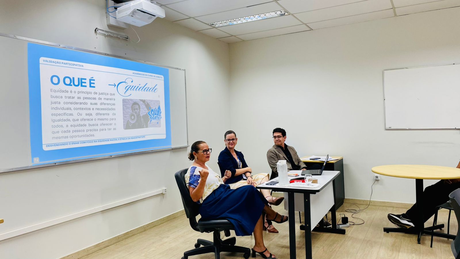 Foto 2: Imagem colorida mostra três pessoas sentadas à frente de uma sala de aula durante uma reunião com estudantes sobre equidade racial no Exame Nacional da Magistratura. Um gráfico circular com dados sobre equidade racial na magistratura brasileira é projetado no quadro branco ao fundo. Algumas pessoas assistem sentadas em cadeiras. Foto 2: Imagem colorida mostra três pessoas sentadas à frente de uma sala de aula durante uma reunião com estudantes sobre equidade racial no Exame Nacional da Magistratura. Um gráfico circular com dados sobre equidade racial na magistratura brasileira é projetado no quadro branco ao fundo. Algumas pessoas assistem sentadas em cadeiras.