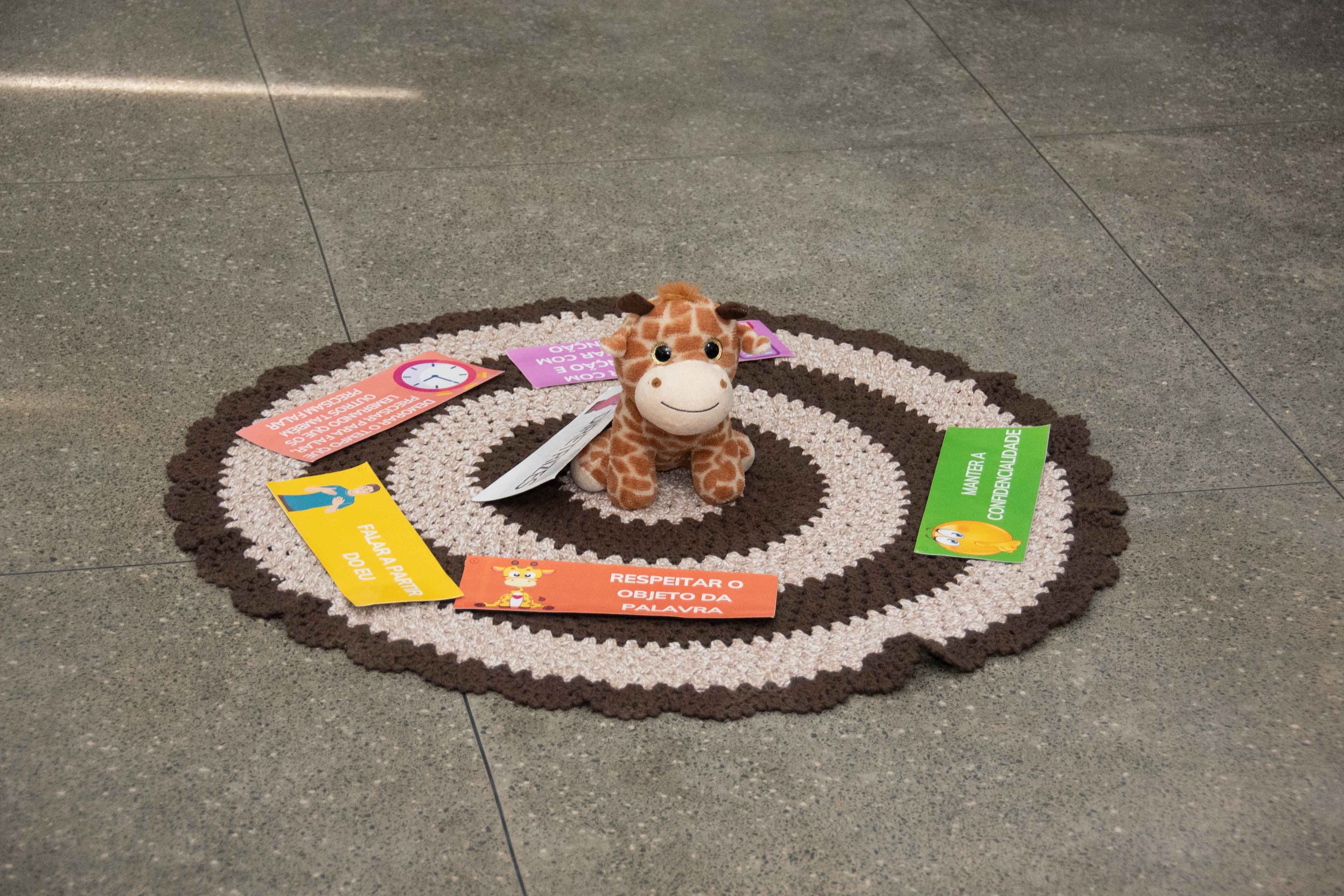 A imagem mostra um tapete redondo de crochê marrom e bege no chão, com uma girafa de pelúcia ao centro. Ao redor da girafa, há cartões coloridos com regras de convivência como “Manter a confidencialidade” e “Não fazer julgamentos”. A imagem mostra um tapete redondo de crochê marrom e bege no chão, com uma girafa de pelúcia ao centro. Ao redor da girafa, há cartões coloridos com regras de convivência como “Manter a confidencialidade” e “Não fazer julgamentos”.