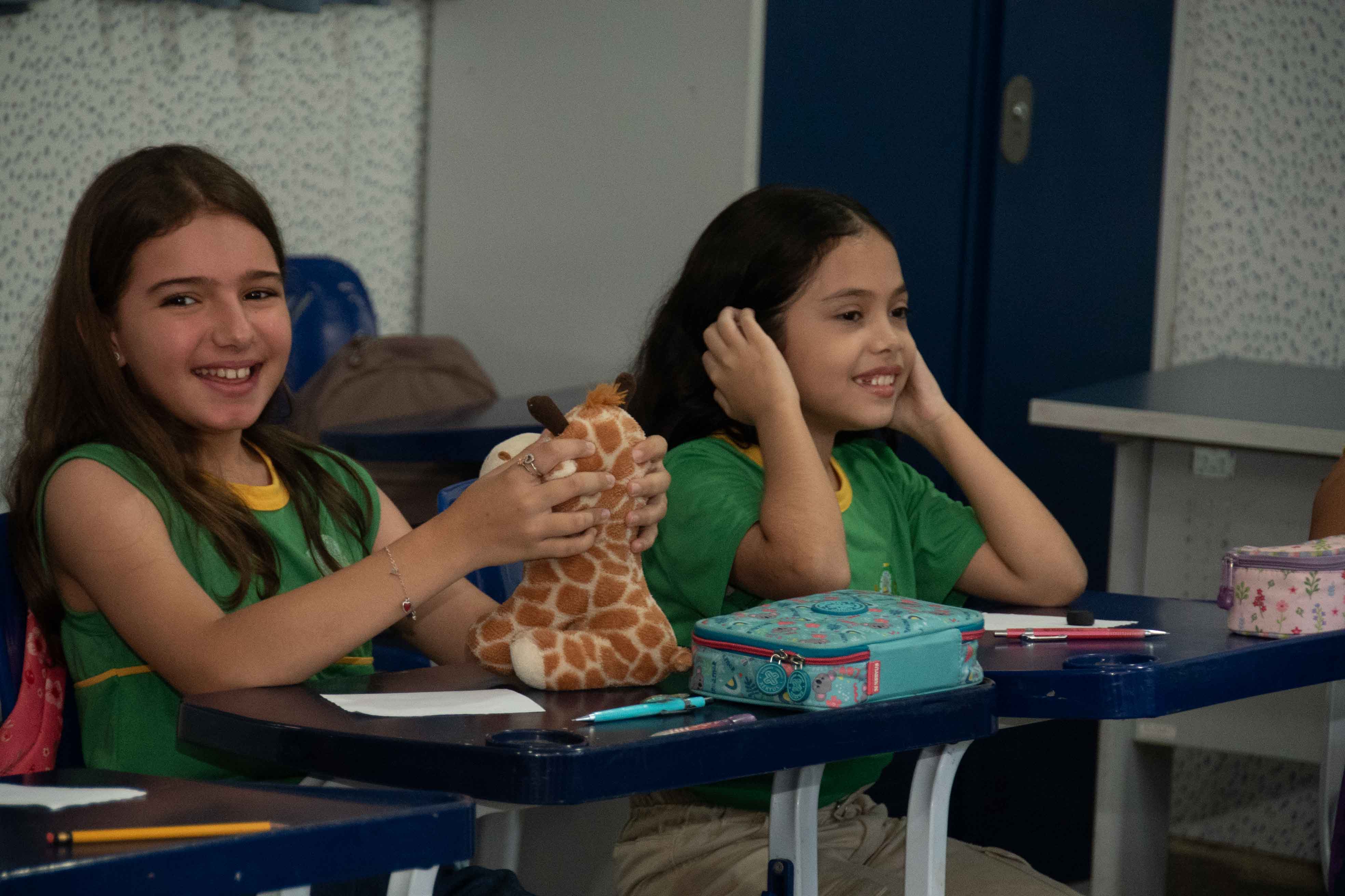Duas meninas, com cerca de 8 a 10 anos, são fotografadas em um momento espontâneo de alegria em sala de aula. Elas usam uniforme verde com detalhes amarelos e estão sentadas lado a lado em uma carteira com tampo azul-escuro. A menina da esquerda sorri para a câmera segurando uma girafa de pelúcia, enquanto a da direita olha para o lado. Sobre a mesa, há materiais escolares como estojo, folhas e canetas. Foto 3: Duas meninas, com cerca de 8 a 10 anos, são fotografadas em um momento espontâneo de alegria em sala de aula. Elas usam uniforme verde com detalhes amarelos e estão sentadas lado a lado em uma carteira com tampo azul-escuro. A menina da esquerda sorri para a câmera segurando uma girafa de pelúcia, enquanto a da direita olha para o lado. Sobre a mesa, há materiais escolares como estojo, folhas e canetas. Foto 3: