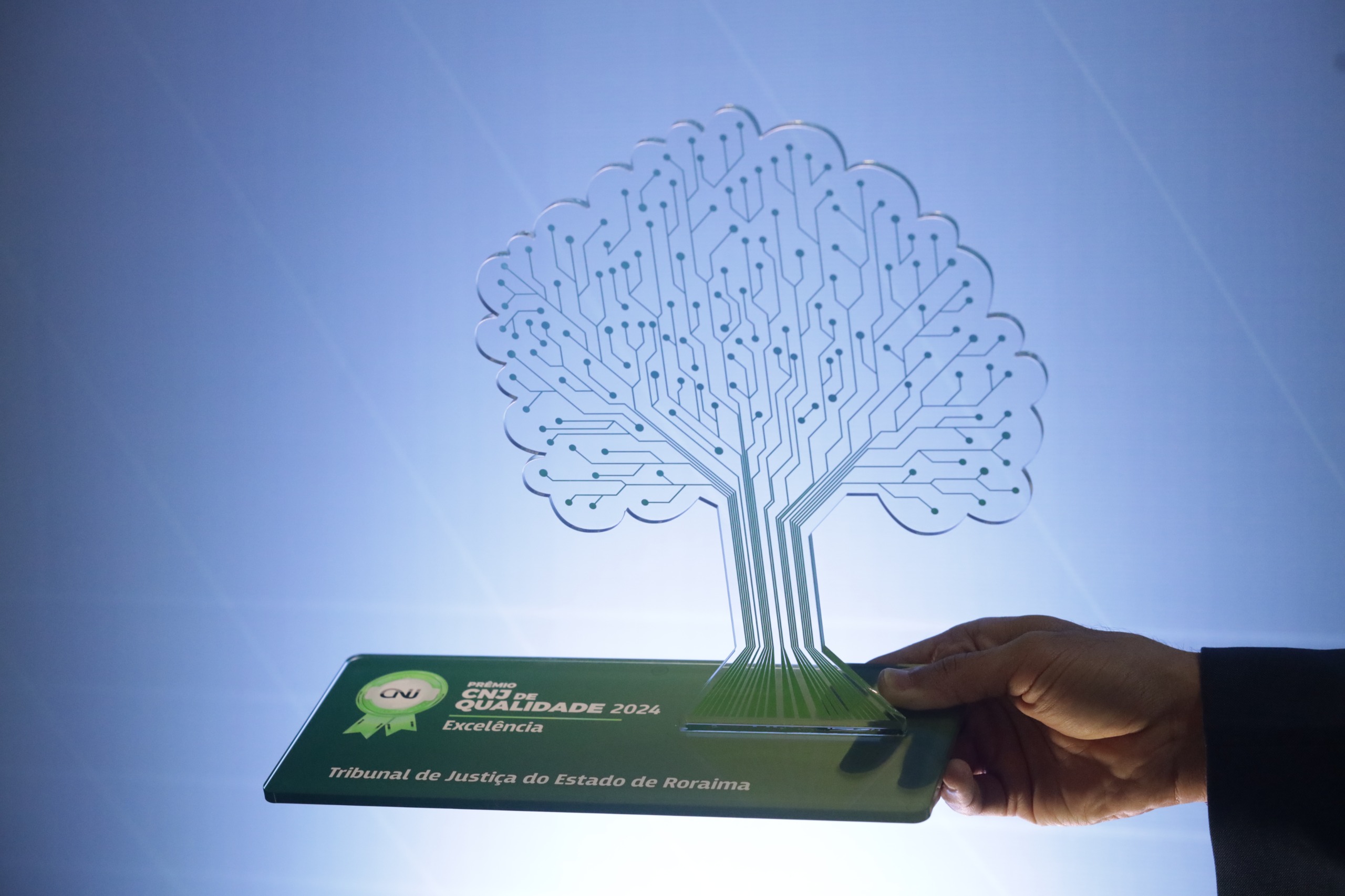 A imagem mostra um troféu transparente em formato de árvore, com linhas que lembram circuitos eletrônicos representando os galhos. A base do troféu é verde, com a inscrição "Prêmio