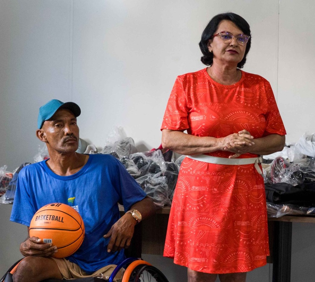 Foto colorida mostra um homem cadeirante com boné azul segurando uma bola de basquete, ao lado da desembargadora Tânia Vasconcelos, que está usando vestido vermelho. Ao fundo, há uma mesa com diversos pares de tênis embalados, sugerindo um evento de doação ou apoio esportivo. Foto colorida mostra um homem cadeirante com boné azul segurando uma bola de basquete, ao lado da desembargadora Tânia Vasconcelos, que está usando vestido vermelho. Ao fundo, há uma mesa com diversos pares de tênis embalados, sugerindo um evento de doação ou apoio esportivo.