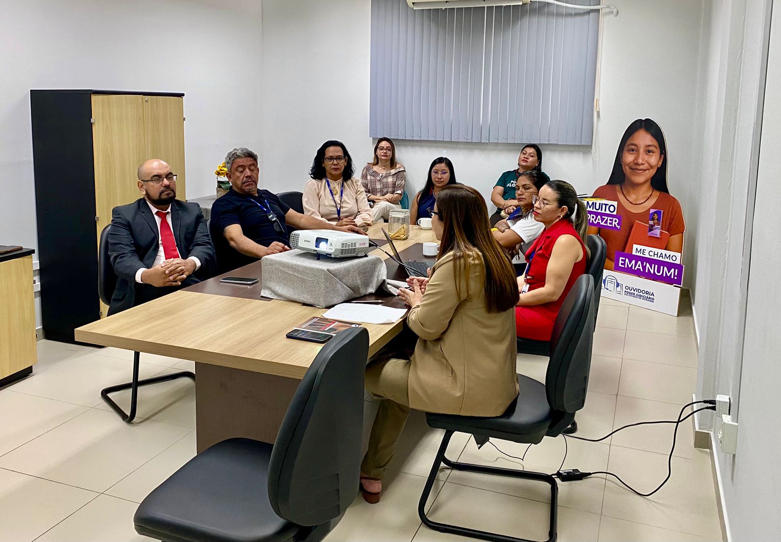 Foto colorida mostra uma reunião em andamento com dez pessoas sentadas em torno de uma mesa de reunião em uma sala do TJRR. No centro da mesa, há um projetor. A maioria dos participantes observa atentamente a mulher que está conduzindo a apresentação. Ao fundo, um totem de papelão em tamanho real exibe a personagem "Ema’num", com a frase: "Muito prazer, me chamo Ema’num!", associada à Ouvidoria do TJRR. Foto colorida mostra uma reunião em andamento com dez pessoas sentadas em torno de uma mesa de reunião em uma sala do TJRR. No centro da mesa, há um projetor. A maioria dos participantes observa atentamente a mulher que está conduzindo a apresentação. Ao fundo, um totem de papelão em tamanho real exibe a personagem "Ema’num", com a frase: "Muito prazer, me chamo Ema’num!", associada à Ouvidoria do TJRR.