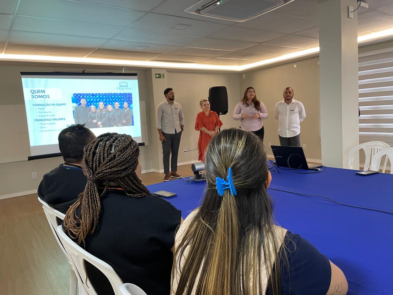 Imagem colorida em ambiente interno com a turma sentada. À esquerda, uma tela grande exibe uma apresentação com texto que inclui "QUEM SOMOS" e "FORMAÇÃO DA EQUIPE" Uma mulher com uma blusa rosa claro está falando. Um grupo de participantes está sentado em uma longa mesa em primeiro plano, de frente para os apresentadores e a tela. A mesa está coberta com um pano azul. Imagem colorida em ambiente interno com a turma sentada. À esquerda, uma tela grande exibe uma apresentação com texto que inclui "QUEM SOMOS" e "FORMAÇÃO DA EQUIPE" Uma mulher com uma blusa rosa claro está falando. Um grupo de participantes está sentado em uma longa mesa em primeiro plano, de frente para os apresentadores e a tela. A mesa está coberta com um pano azul.