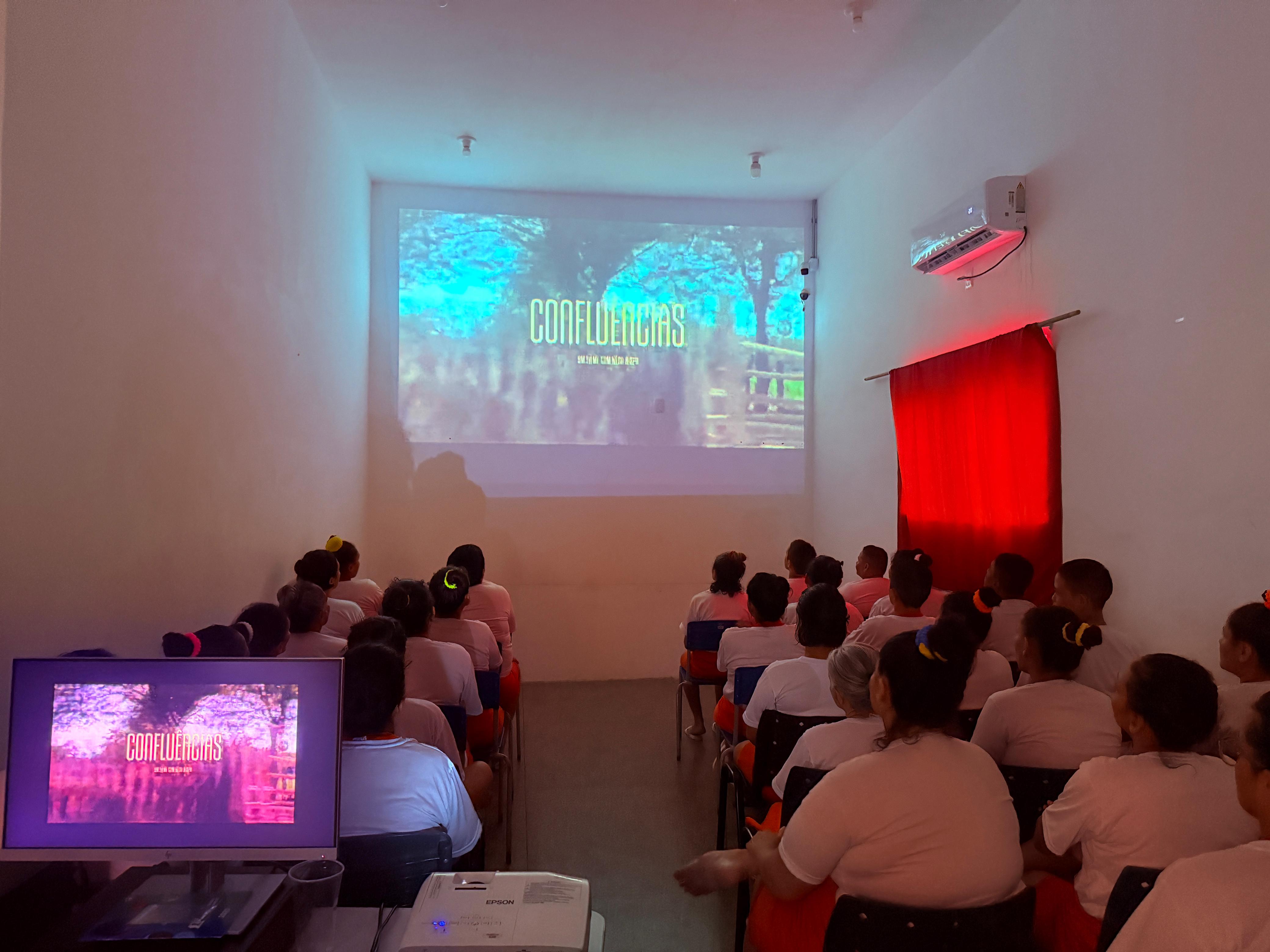 Foto colorida mostra um grupo de mulheres assistindo a uma sessão de cinema em uma sala com luz baixa. Elas vestem roupas claras e estão voltadas para uma tela onde é exibido um filme. À frente, um projetor e um monitor duplicam a imagem. A sala tem paredes brancas e uma cortina vermelha. Foto colorida mostra um grupo de mulheres assistindo a uma sessão de cinema em uma sala com luz baixa. Elas vestem roupas claras e estão voltadas para uma tela onde é exibido um filme. À frente, um projetor e um monitor duplicam a imagem. A sala tem paredes brancas e uma cortina vermelha.