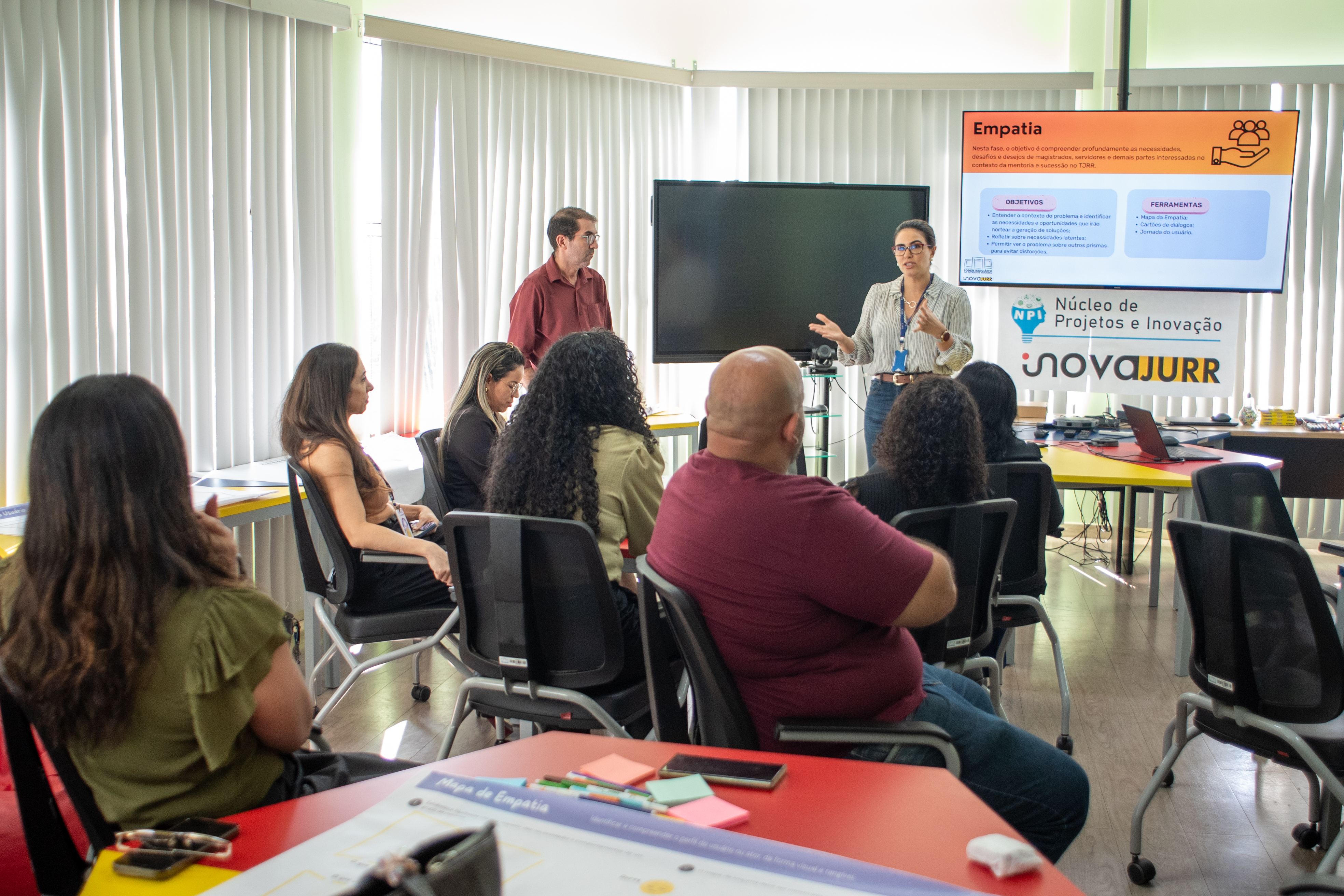 Foto colorida mostra uma mulher apresentando uma palestra para um grupo de pessoas sentadas em uma sala. Foto colorida mostra uma mulher apresentando uma palestra para um grupo de pessoas sentadas em uma sala.