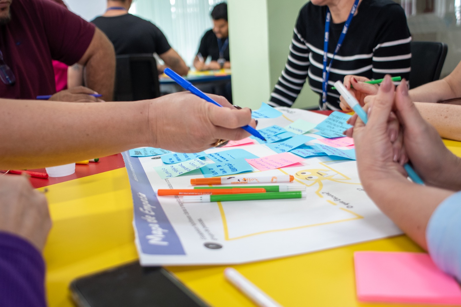 Foto colorida mostra pessoas participando de uma atividade em grupo, escrevendo em papéis coloridos sobre uma cartolina com a frase “Mapa da Empatia”. Há várias canetas coloridas e post-its com anotações sobre a mesa. A imagem destaca mãos em ação e foco colaborativo. Foto colorida mostra pessoas participando de uma atividade em grupo, escrevendo em papéis coloridos sobre uma cartolina com a frase “Mapa da Empatia”. Há várias canetas coloridas e post-its com anotações sobre a mesa. A imagem destaca mãos em ação e foco colaborativo.