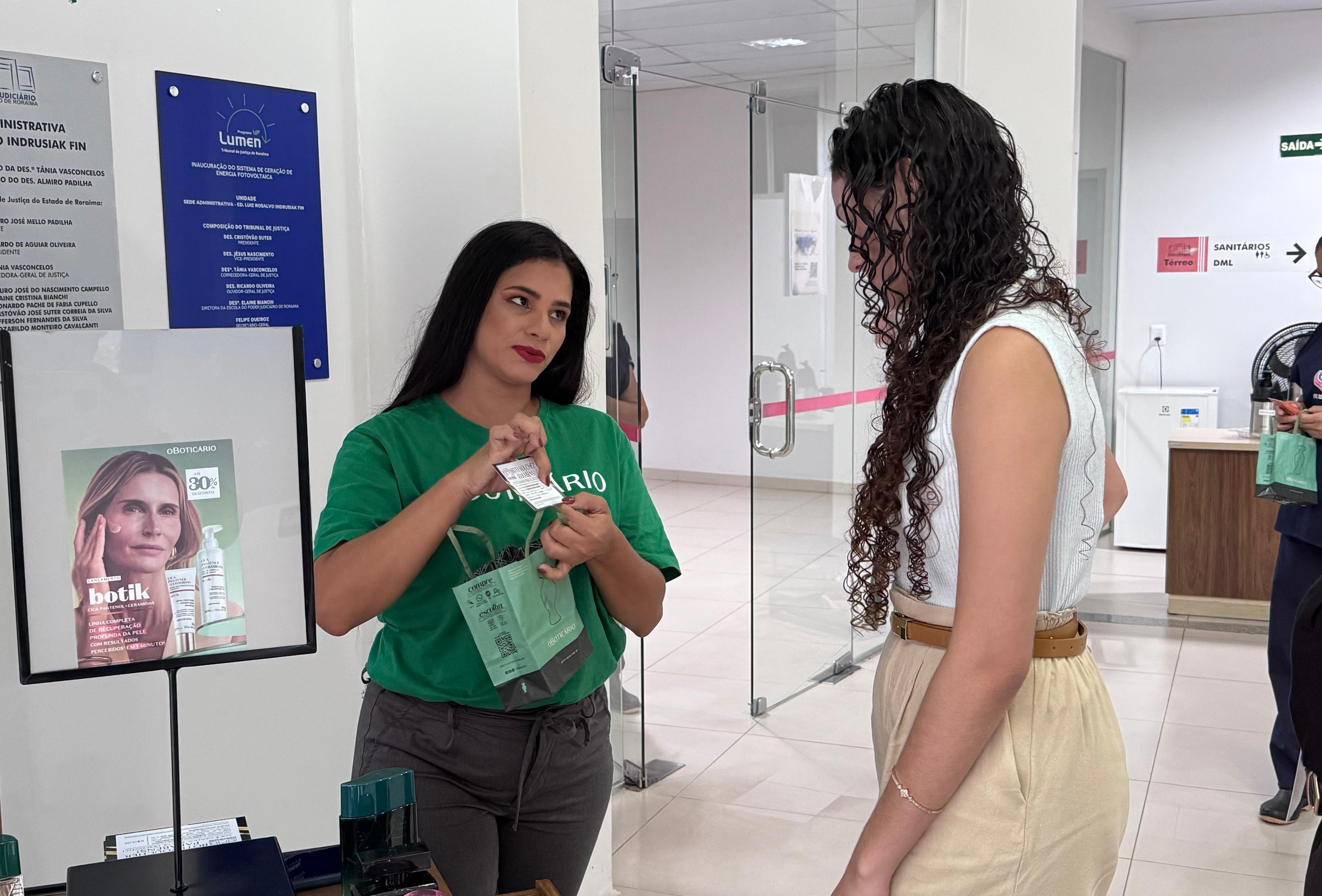 Imagem colorida mostra duas mulheres em um ambiente interno. A mulher à esquerda, vestindo uma camiseta verde da marca O Boticário, está segurando uma sacola de presente verde e um pequeno cartão, enquanto fala com a mulher à direita. A mulher à direita usa uma blusa clara e uma calça bege. Ao fundo, há uma parede branca com placas informativas, uma porta de vidro e um cartaz promocional de produtos de beleza.
