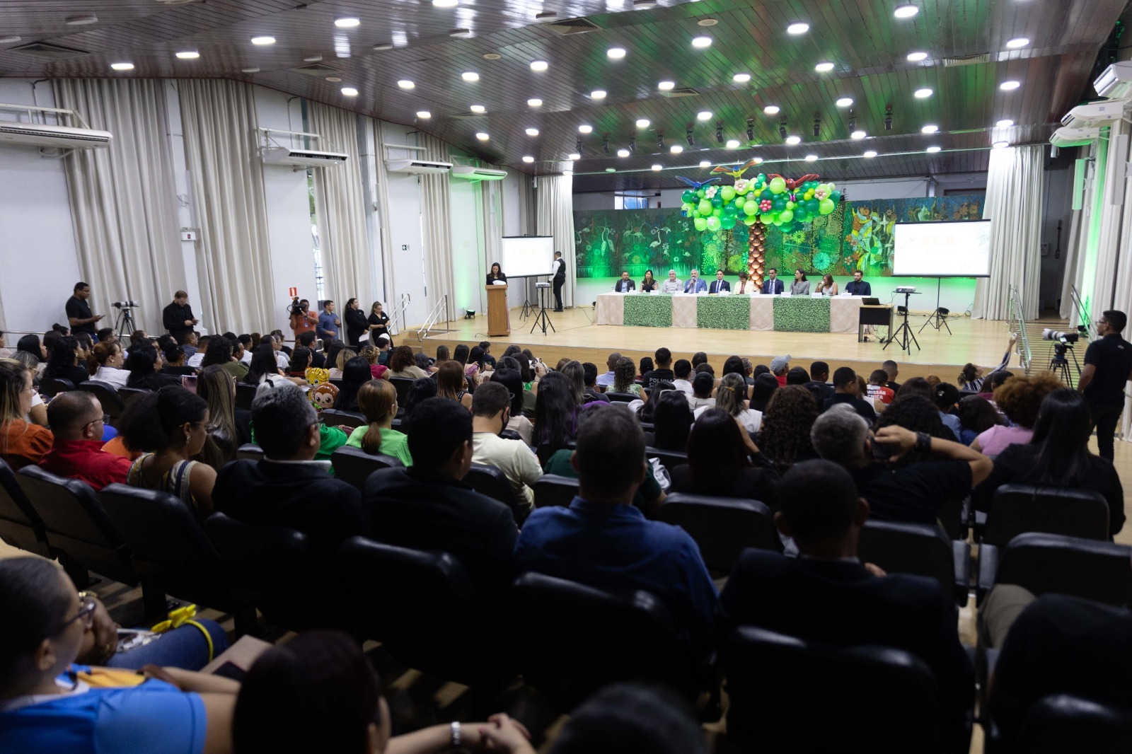 Imagem colorida mostra uma vista panorâmica de um grande auditório. Uma palestrante está em pé no palco, à esquerda, com um púlpito e uma tela de projeção. No centro do palco, há uma longa mesa com vários participantes sentados. Uma decoração com balões verdes em formato de árvore está no palco. A plateia está sentada em várias fileiras de cadeiras escuras, de costas para a câmera, assistindo à apresentação. As paredes laterais são cobertas por cortinas claras.