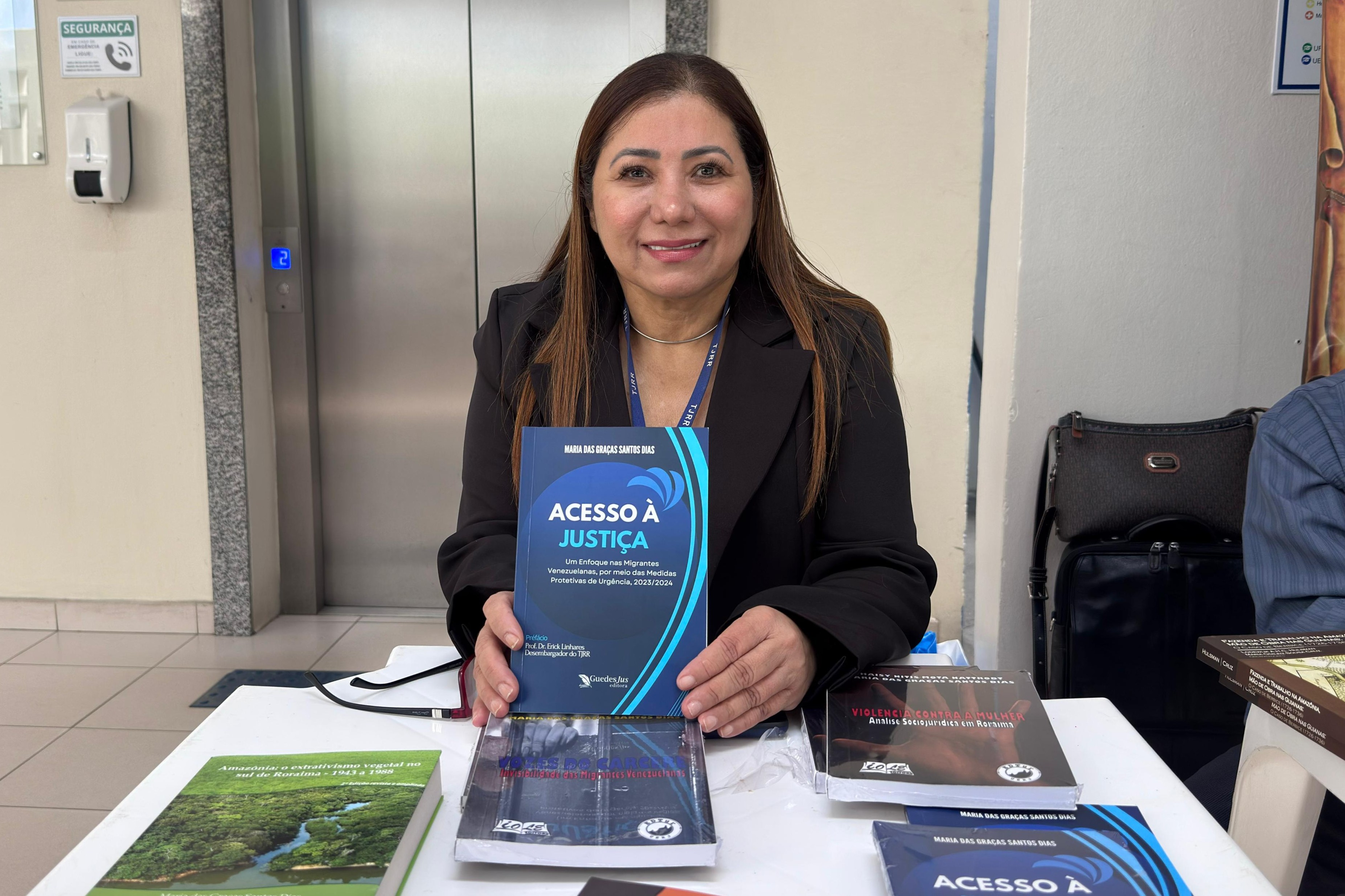 Foto colorida mostra uma mulher de cabelos lisos castanhos, trajando blazer preto e crachá institucional do TJRR, sorri enquanto segura o livro "Acesso à Justiça", de sua autoria — Maria das Graças Santos Dias. À sua frente, sobre a mesa, estão outros títulos também voltados à temática dos direitos humanos e justiça. Ao fundo, há um elevador de portas metálicas e paredes claras. A imagem foi registrada durante a Feira do Livro realizada no Tribunal de Justiça de Roraima.