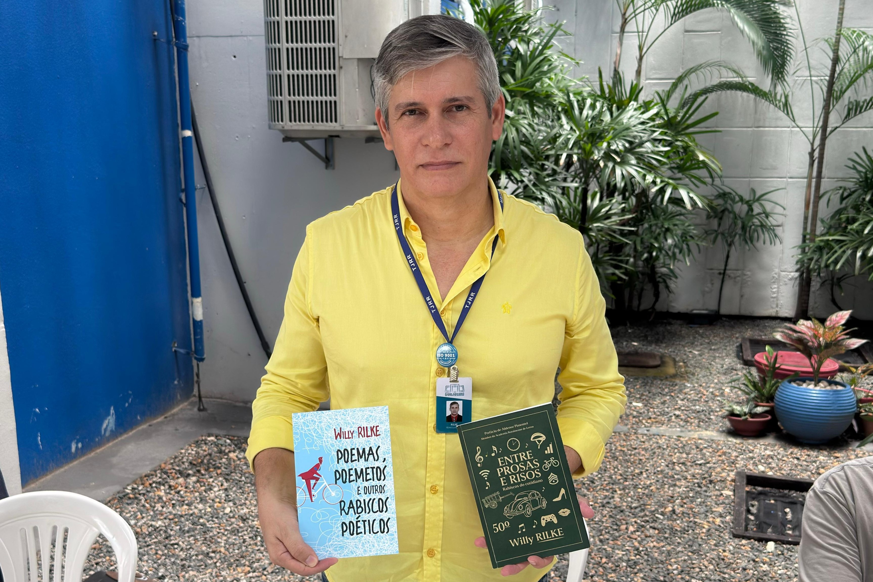 Foto colorida mostra um homem de cabelos grisalhos vestindo uma camisa amarela e crachá do Tribunal de Justiça de Roraima. Ele segura dois livros: à esquerda, “Poemas, Poemetos e outros Rabiscos Poéticos”, com capa azul clara e ilustração de uma pessoa andando de bicicleta; à direita, “Entre Prosas e Risos”, de capa verde escura com ícones dourados e o nome do autor Willy Rilke.  Foto 5: Foto colorida mostra uma mulher de cabelos lisos castanhos, trajando blazer preto e crach