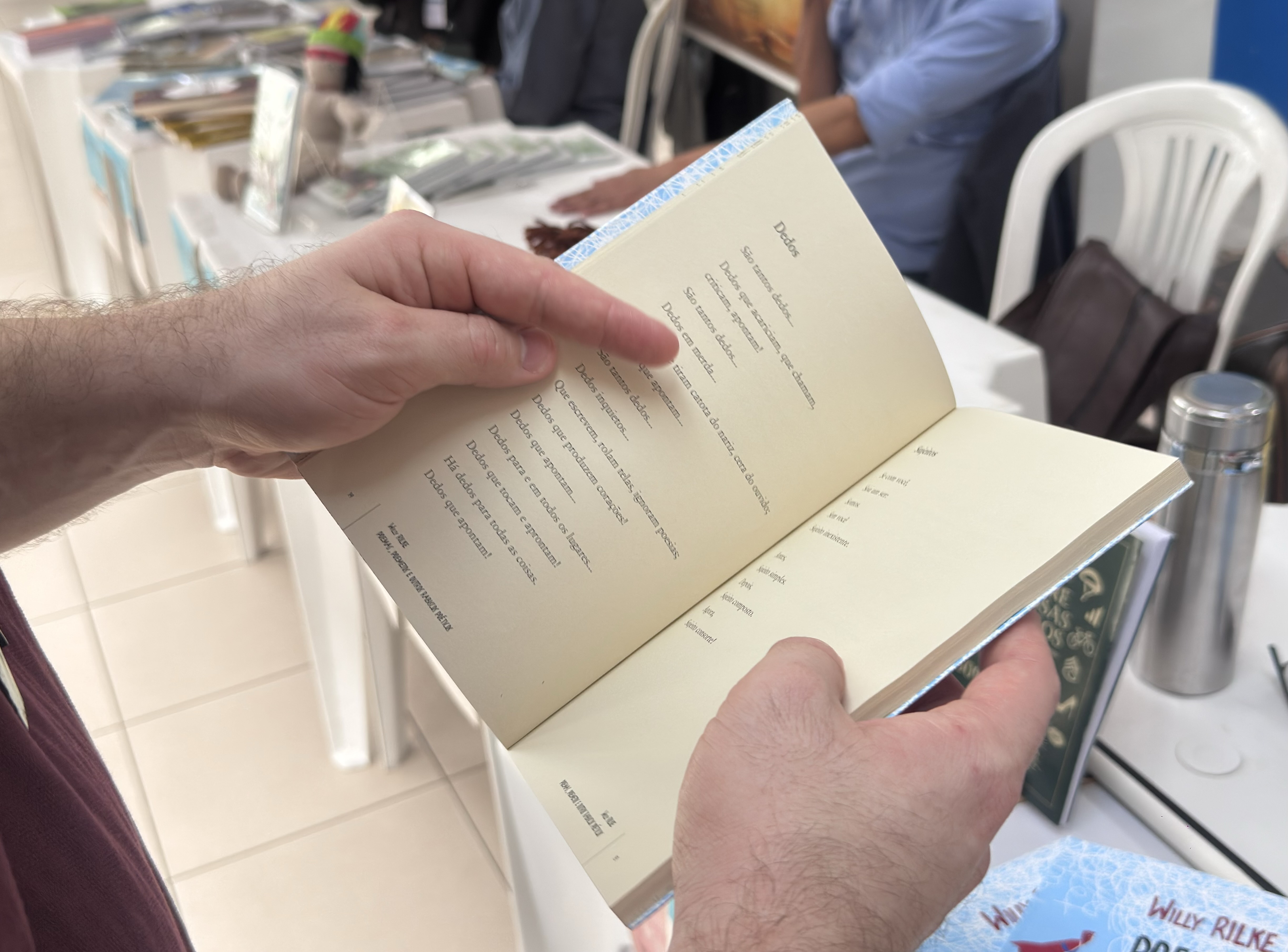 Foto colorida de uma pessoa folheando um livro em um ambiente de feira ou exposição literária. A imagem mostra de perto as mãos da pessoa, que segura um livro aberto com páginas em tom amarelado. Ao fundo, há mesas com livros expostos, cadeiras brancas e pessoas sentadas. 