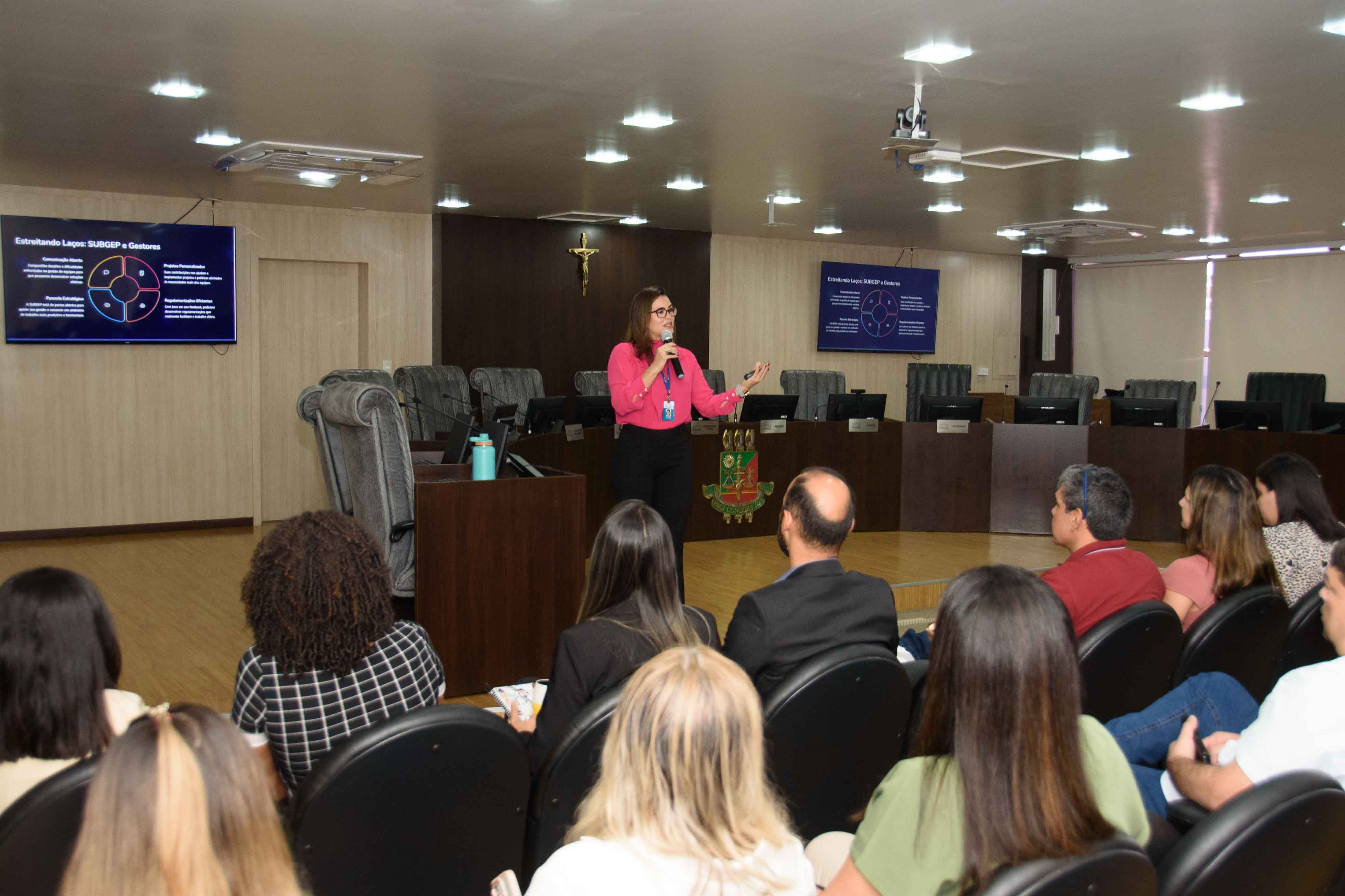 Imagem colorida mostra uma mulher morena de óculos, vestindo uma blusa rosa e calça preta, em pé ao lado de um pódio de madeira, falando e gesticulando com a mão direita. Ao fundo, duas telas exibem slides de apresentação com texto e diagramas, e uma plateia atenta preenche uma sala formal com cadeiras escuras e iluminação de teto. Imagem colorida mostra uma mulher morena de óculos, vestindo uma blusa rosa e calça preta, em pé ao lado de um pódio de madeira, falando e gesticulando com a mão direita. Ao fundo, duas telas exibem slides de apresentação com texto e diagramas, e uma plateia atenta preenche uma sala formal com cadeiras escuras e iluminação de teto.