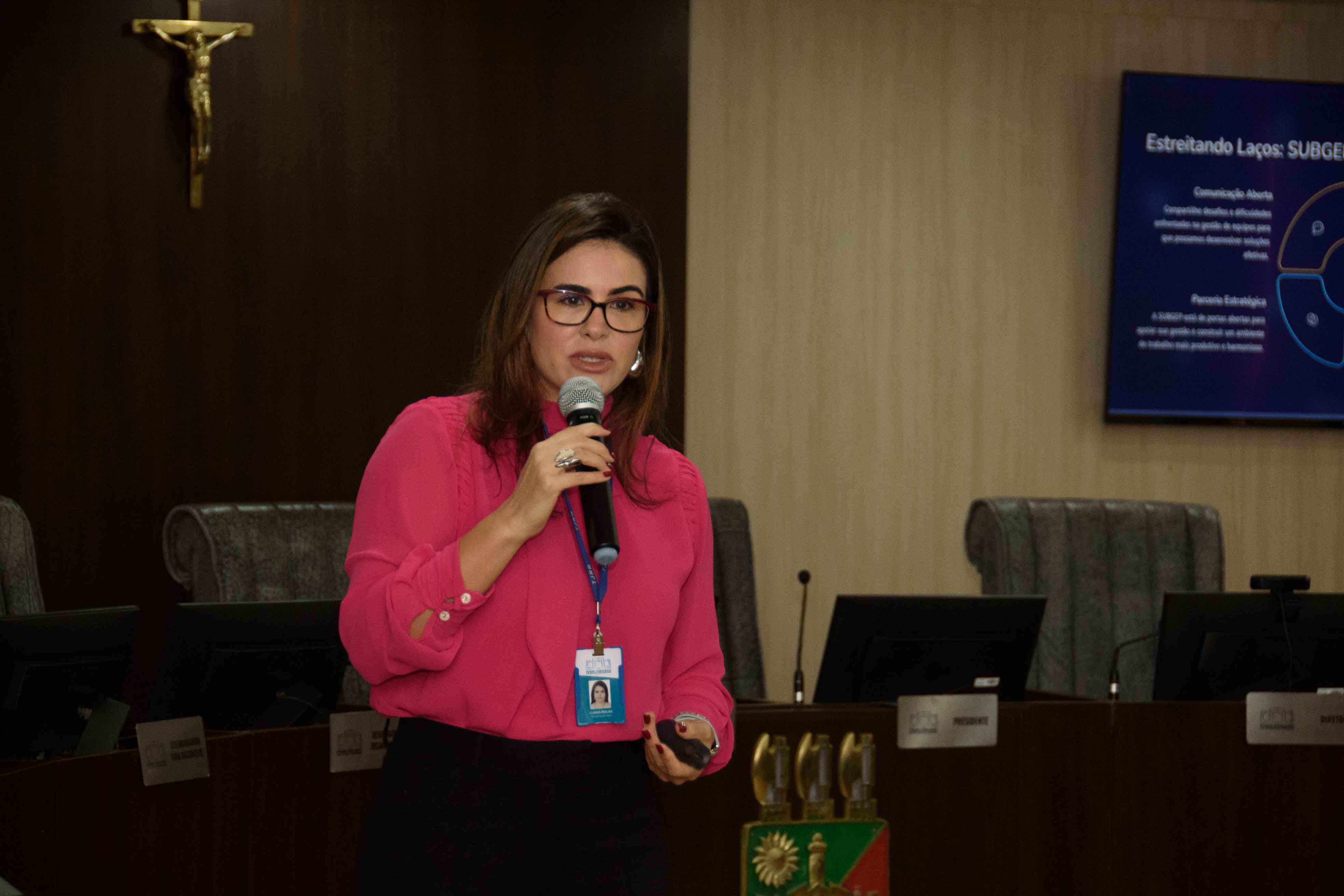 Imagem colorida mostra uma mulher morena de óculos, vestindo uma blusa rosa e calça preta, falando em um microfone em um pódio de madeira em frente a uma plateia atenta. Ao fundo, uma tela exibe slides de apresentação com texto e diagramas, em uma sala formal com cadeiras escuras e iluminação de teto. Imagem colorida mostra uma mulher morena de óculos, vestindo uma blusa rosa e calça preta, falando em um microfone em um pódio de madeira em frente a uma plateia atenta. Ao fundo, uma tela exibe slides de apresentação com texto e diagramas, em uma sala formal com cadeiras escuras e iluminação de teto.