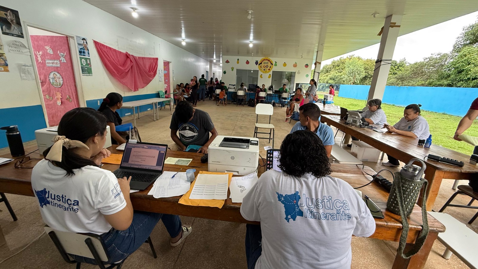 Foto colorida mostra ação da Justiça Itinerante em uma área coberta, com várias mesas de atendimento. Pessoas trabalham em laptops e realizam cadastros, enquanto moradores aguardam sentados ao fundo. Os atendentes vestem camisetas brancas com a logo “Justiça Itinerante” nas costas. Foto colorida mostra ação da Justiça Itinerante em uma área coberta, com várias mesas de atendimento. Pessoas trabalham em laptops e realizam cadastros, enquanto moradores aguardam sentados ao fundo. Os atendentes vestem camisetas brancas com a logo “Justiça Itinerante” nas costas.