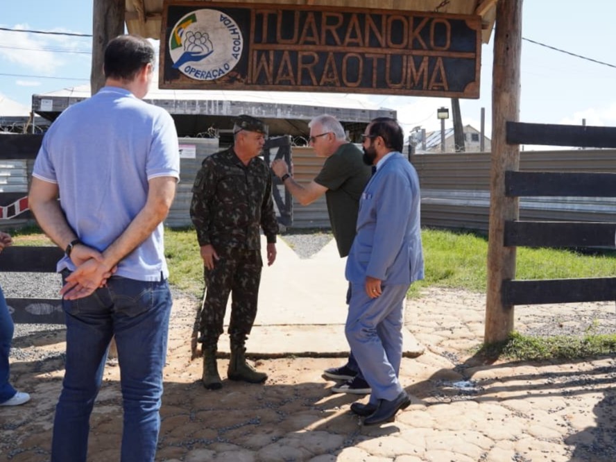 imagem colorida mostra o ministro, o presidente do TJRR, o desembargador Cristovão Súter e um militar em uma área externa, ao ar livre. Ao fundo, há um portão de madeira com uma placa grande e marrom, com o texto "ACOLHIDA ITURANOKO IWARAOTUMA" e "OPERAÇÃO ACOLHIDA". imagem colorida mostra o ministro, o presidente do TJRR, o desembargador Cristovão Súter e um militar em uma área externa, ao ar livre. Ao fundo, há um portão de madeira com uma placa grande e marrom, com o texto "ACOLHIDA ITURANOKO IWARAOTUMA" e "OPERAÇÃO ACOLHIDA".