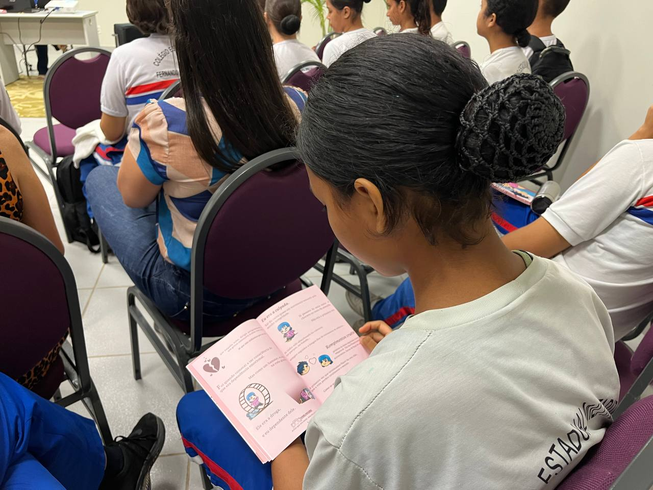 A foto mostra uma aluna sentada, de perfil, folheando um livreto educativo de cor rosa. O conteúdo do livreto parece tratar de relacionamentos afetivos entre adolescentes, com ilustrações e frases curtas. A estudante veste camiseta branca com o nome da escola nas costas. Ao fundo, outros alunos também estão sentados assistindo à palestra. Assim como na primeira imagem, há uma faixa azul com os dizeres: A foto mostra uma aluna sentada, de perfil, folheando um livreto educativo de cor rosa. O conteúdo do livreto parece tratar de relacionamentos afetivos entre adolescentes, com ilustrações e frases curtas. A estudante veste camiseta branca com o nome da escola nas costas. Ao fundo, outros alunos também estão sentados assistindo à palestra. Assim como na primeira imagem, há uma faixa azul com os dizeres: