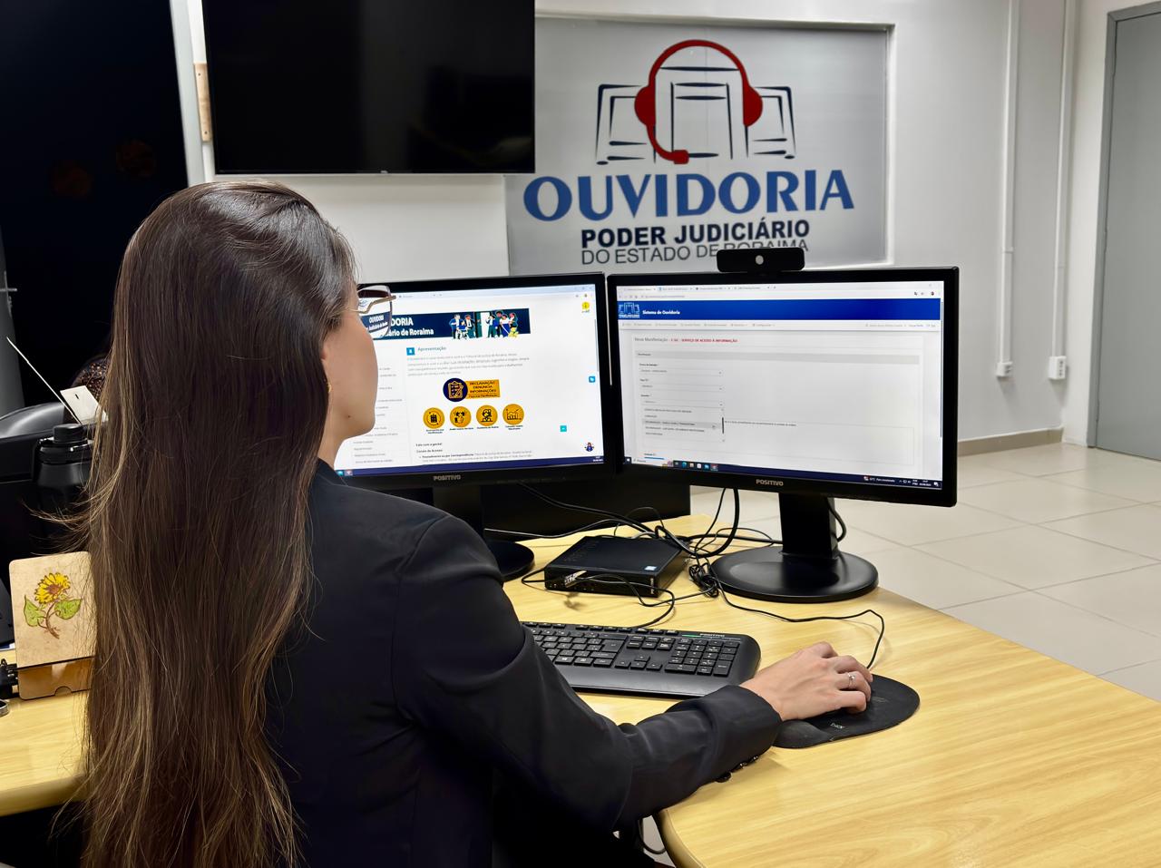 Foto colorida mostra uma mulher trabalhando em dois monitores na Ouvidoria do Poder Judiciário de Roraima. O monitor esquerdo exibe a página principal da ouvidoria, enquanto o direito mostra um formulário online. No fundo, um banner reforça a identidade visual da "OUVIDORIA PODER JUDICIÁRIO DO ESTADO DE RORAIMA". Foto colorida mostra uma mulher trabalhando em dois monitores na Ouvidoria do Poder Judiciário de Roraima. O monitor esquerdo exibe a página principal da ouvidoria, enquanto o direito mostra um formulário online. No fundo, um banner reforça a identidade visual da "OUVIDORIA PODER JUDICIÁRIO DO ESTADO DE RORAIMA".