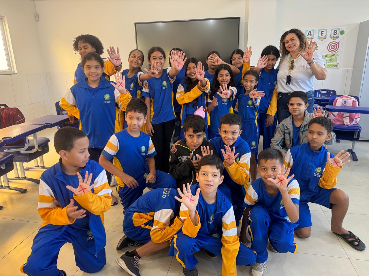 Imagem colorida mostra um grupo de crianças e uma mulher adulta posa em uma sala de aula. As crianças, usando uniformes azuis e amarelos, estão em pé ou agachadas e levantam as mãos. A mulher adulta, à direita, também levanta as mãos com o desenho. Ao fundo, uma lousa digital e cartazes. Imagem colorida mostra um grupo de crianças e uma mulher adulta posa em uma sala de aula. As crianças, usando uniformes azuis e amarelos, estão em pé ou agachadas e levantam as mãos. A mulher adulta, à direita, também levanta as mãos com o desenho. Ao fundo, uma lousa digital e cartazes.