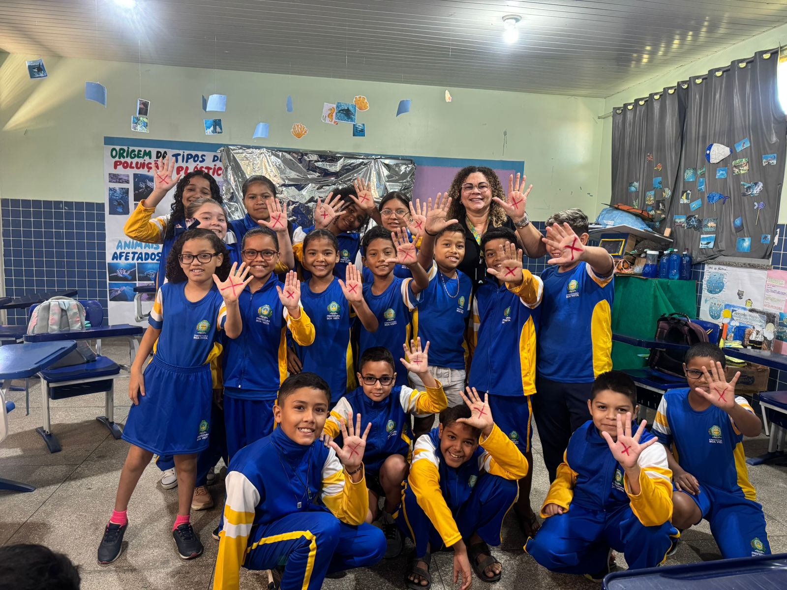 : Imagem colorida mostra um grupo de crianças e uma mulher adulta posam para a foto em uma sala de aula. As crianças, vestindo uniformes azuis e amarelos, estão em diferentes posições (algumas em pé, outras agachadas) e todas levantam as mãos. Ao fundo, há cartazes com informações sobre poluição e animais marinhos, mesas e cadeiras. : Imagem colorida mostra um grupo de crianças e uma mulher adulta posam para a foto em uma sala de aula. As crianças, vestindo uniformes azuis e amarelos, estão em diferentes posições (algumas em pé, outras agachadas) e todas levantam as mãos. Ao fundo, há cartazes com informações sobre poluição e animais marinhos, mesas e cadeiras.