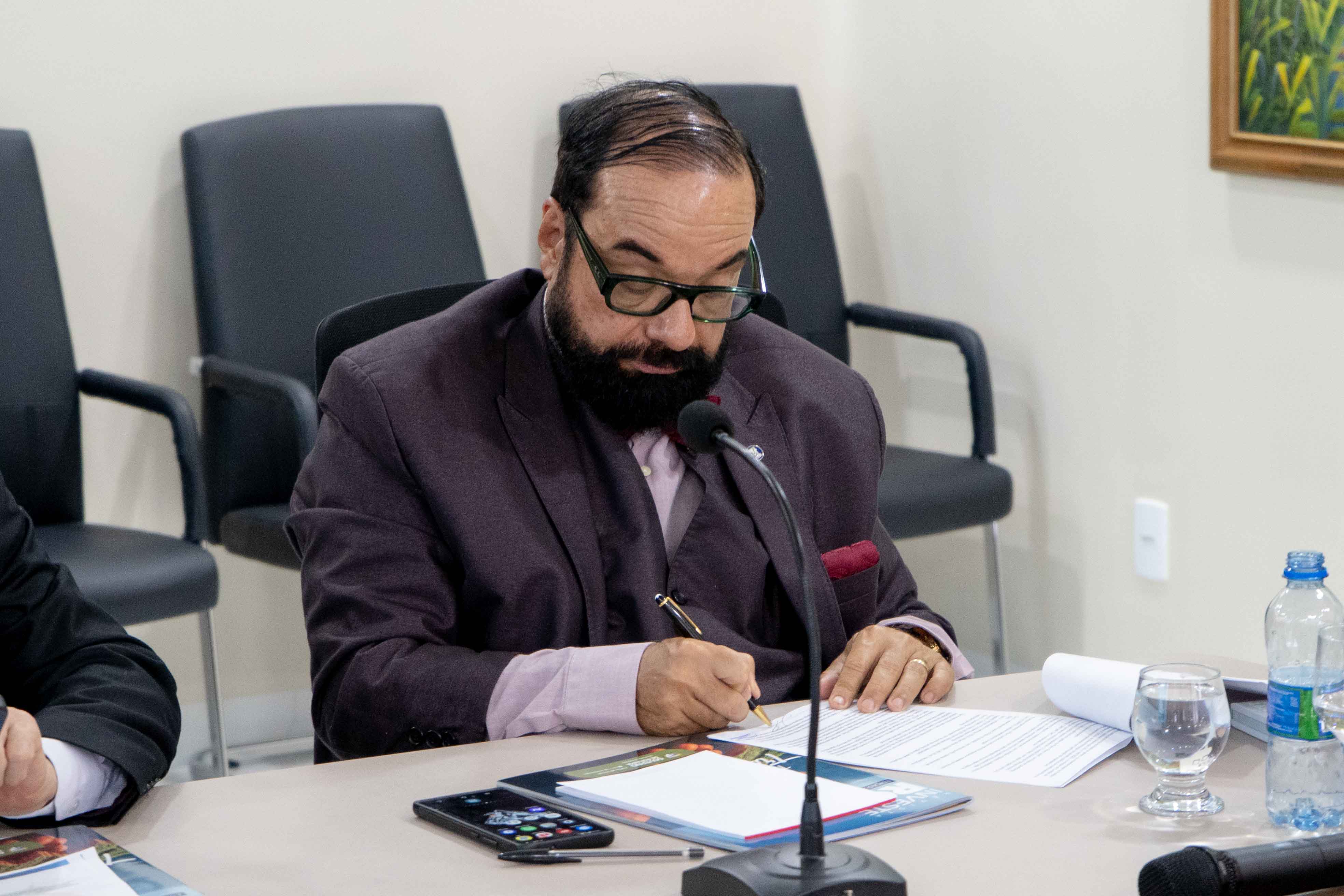 Imagem colorida mostra o Presidente do Tribunal de Justiça de Roraima, Desembargador Leonardo Cupello, sentado em uma mesa de reunião, assinando um documento. Há um microfone, um celular, copos e uma garrafa de água na mesa. Ao fundo, cadeiras de escritório e um quadro na parede são visíveis. Imagem colorida mostra o Presidente do Tribunal de Justiça de Roraima, Desembargador Leonardo Cupello, sentado em uma mesa de reunião, assinando um documento. Há um microfone, um celular, copos e uma garrafa de água na mesa. Ao fundo, cadeiras de escritório e um quadro na parede são visíveis.