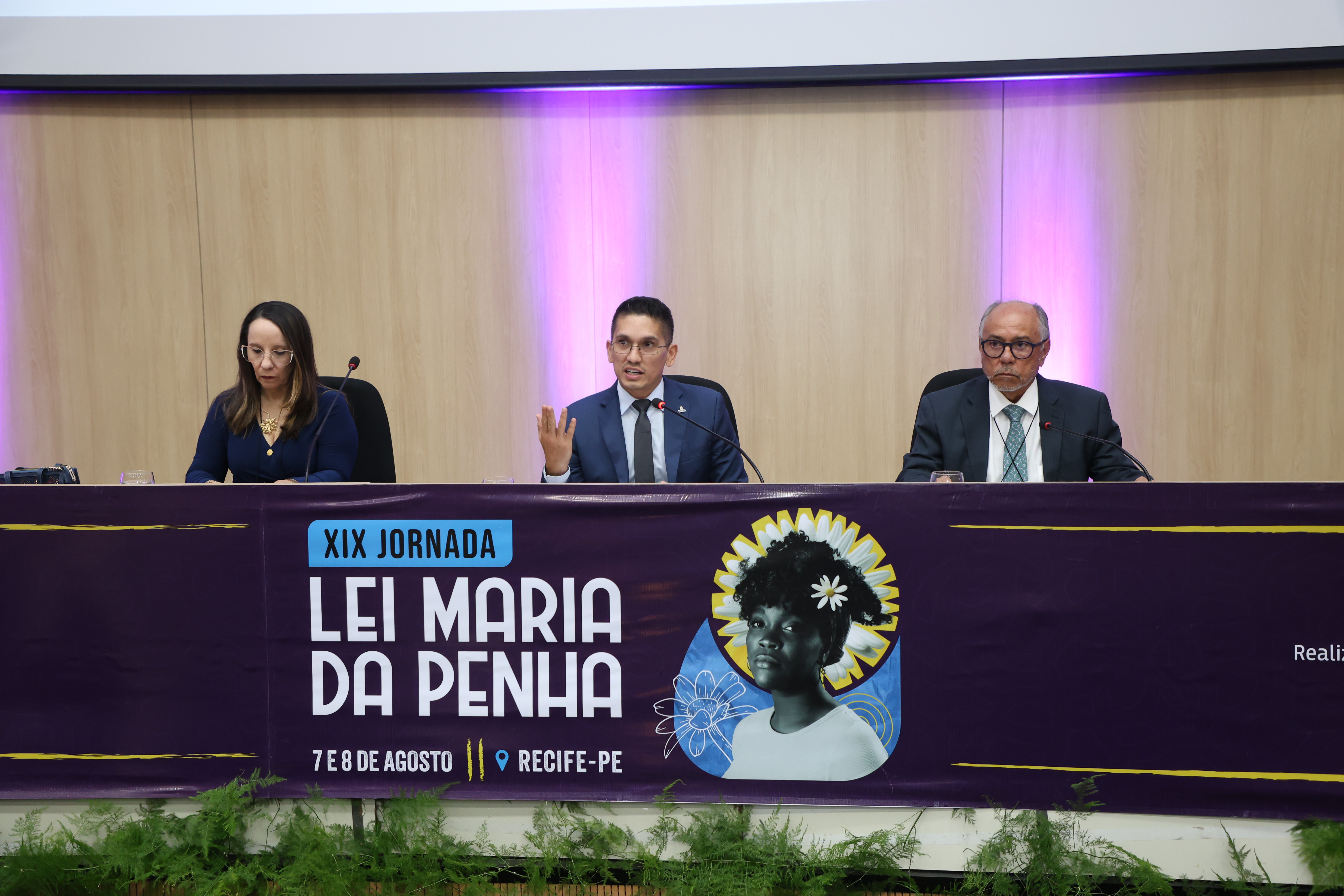 A imagem mostra uma mesa de palestrantes em um evento. No centro, um banner roxo com a inscrição "XIX JORNADA LEI MARIA DA PENHA". A data e o local do evento estão indicados como "7 E 8 DE AGOSTO" e "RECIFE-PE". Ao lado do texto, há uma ilustração de uma mulher negra com um adorno na cabeça. Na mesa, há três pessoas: uma mulher de óculos à esquerda, o Juiz titular da 2ª Vara da Infância e Juventude, Marcelo de Oliveira falando ao microfone e gesticulando, e um homem mais velho de óculos à direita. A imagem mostra uma mesa de palestrantes em um evento. No centro, um banner roxo com a inscrição "XIX JORNADA LEI MARIA DA PENHA". A data e o local do evento estão indicados como "7 E 8 DE AGOSTO" e "RECIFE-PE". Ao lado do texto, há uma ilustração de uma mulher negra com um adorno na cabeça. Na mesa, há três pessoas: uma mulher de óculos à esquerda, o Juiz titular da 2ª Vara da Infância e Juventude, Marcelo de Oliveira falando ao microfone e gesticulando, e um homem mais velho de óculos à direita.
