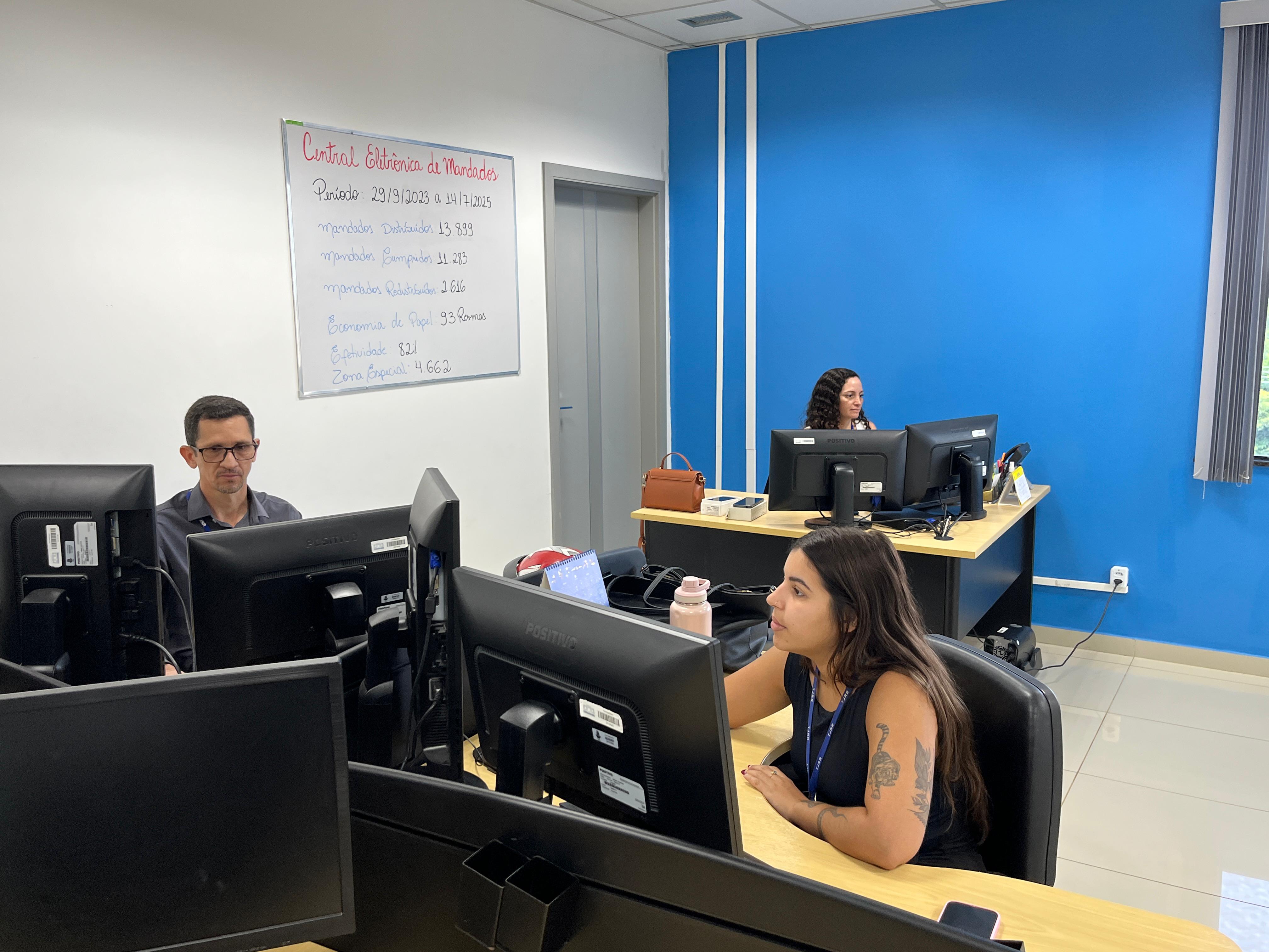 A imagem mostra um ambiente de escritório com três pessoas trabalhando em frente a computadores de mesa.