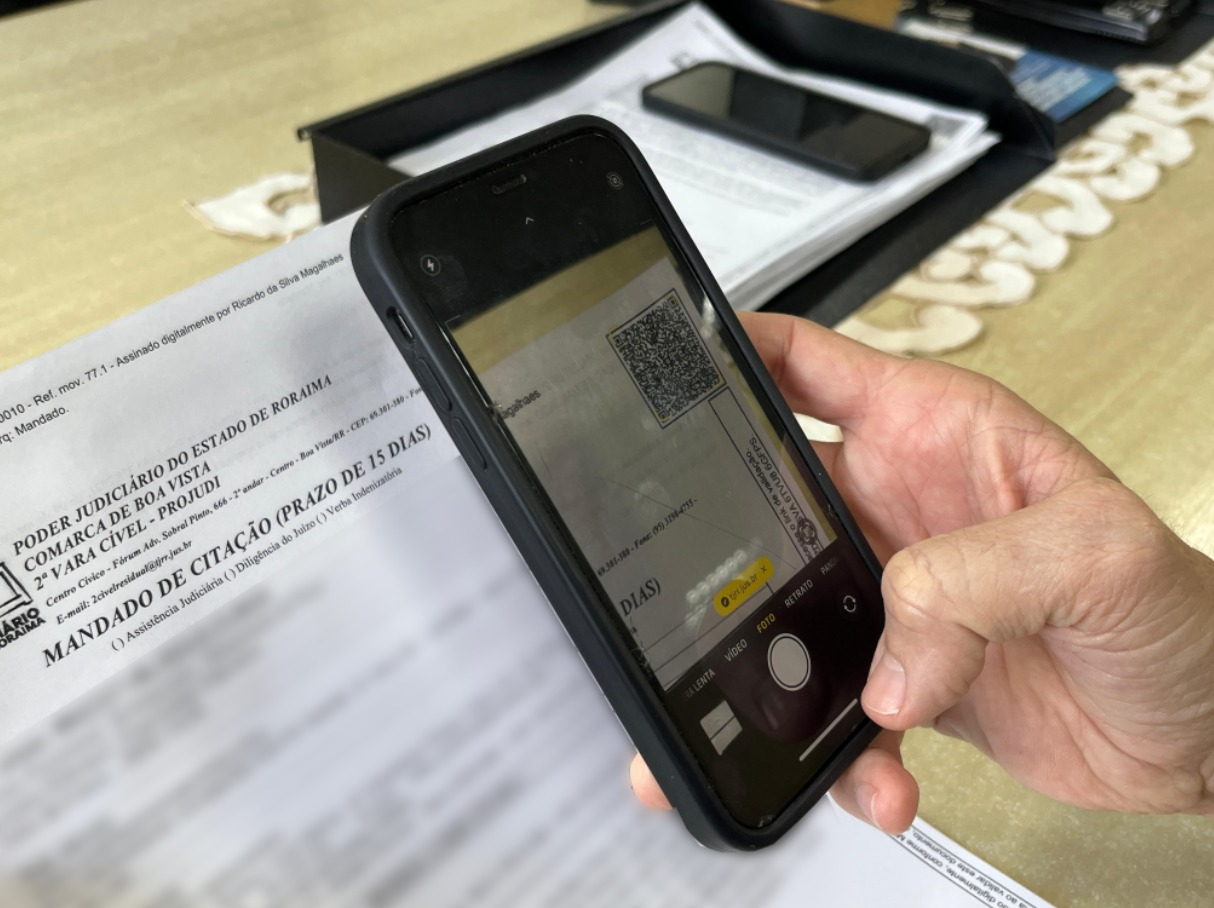 A imagem mostra uma mão segurando um smartphone na posição vertical, com a câmera aberta no modo “Foto”. Na tela do celular, vê-se o enquadramento de um documento impresso com um QR code visível. No topo do papel, é possível ler o cabeçalho “PODER JUDICIÁRIO DO ESTADO DE RORAIMA”, seguido por informações da “2ª Vara Cível – PROJUDI” e o título “MANDADO DE CITAÇÃO (PRAZO DE 15 DIAS)”.