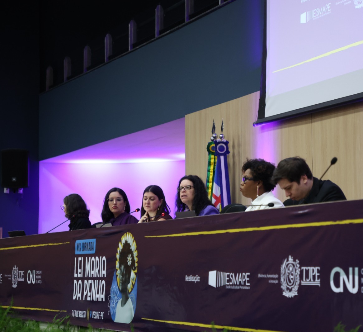 A imagem mostra um painel de palestrantes, em uma mesa com um banner roxo à frente. A inscrição no banner indica "XIX JORNADA LEI MARIA DA PENHA", com a data e o local "7 E 8 DE AGOSTO | RECIFE-PE". Na mesa, há seis pessoas sentadas dentre elas está a juíza coordenadora estadual da Mulher em Situação de Violência Doméstica e Familiar do TJRR, Suelen Alves. No fundo, há um telão com o logotipo do evento e outras informações. A imagem mostra um painel de palestrantes, em uma mesa com um banner roxo à frente. A inscrição no banner indica "XIX JORNADA LEI MARIA DA PENHA", com a data e o local "7 E 8 DE AGOSTO | RECIFE-PE". Na mesa, há seis pessoas sentadas dentre elas está a juíza coordenadora estadual da Mulher em Situação de Violência Doméstica e Familiar do TJRR, Suelen Alves. No fundo, há um telão com o logotipo do evento e outras informações.