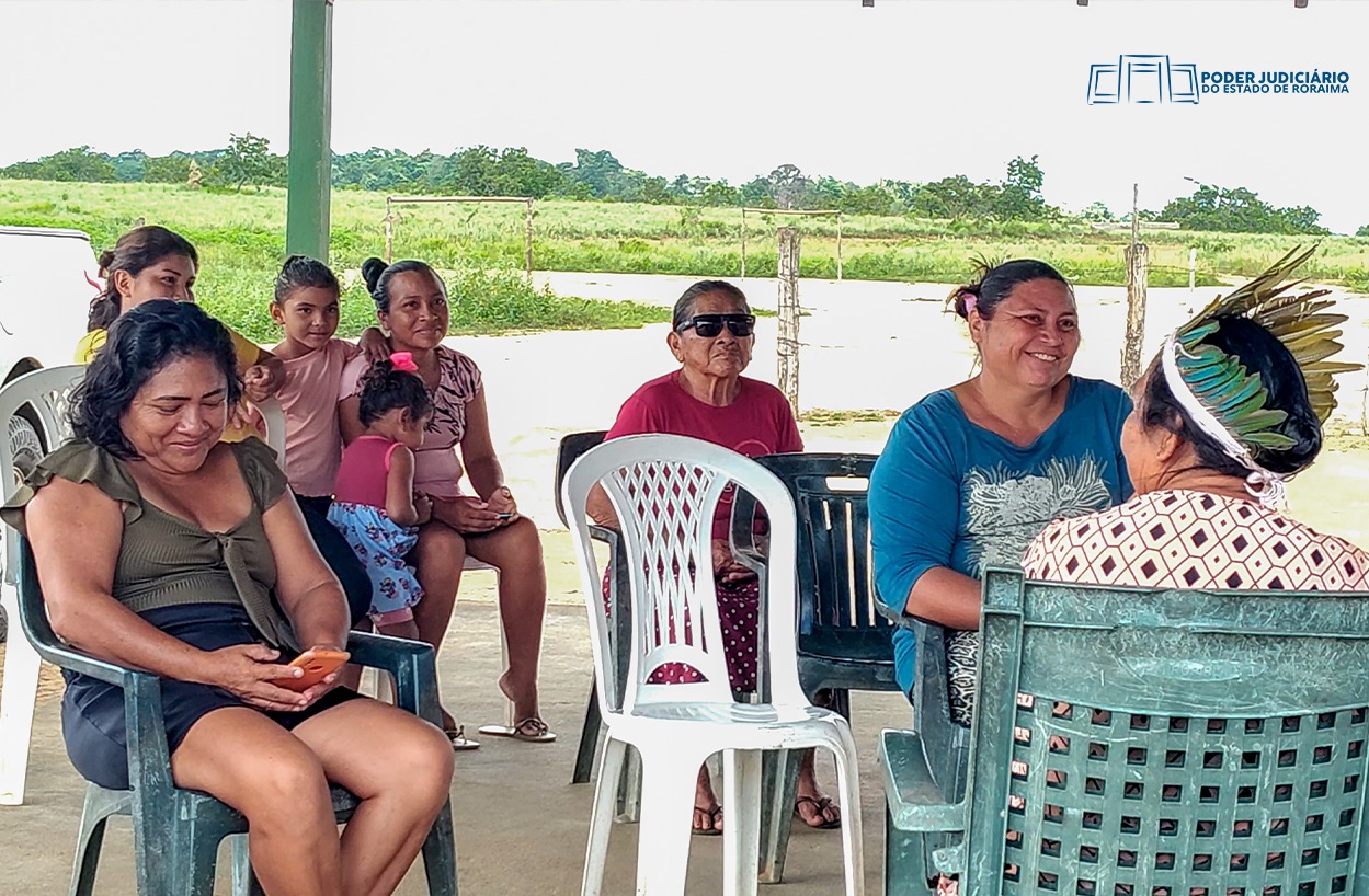 A imagem mostra um grupo de pessoas sentadas em cadeiras de plástico, em um ambiente que parece ser externo, com vegetação ao fundo. Há crianças e adultos, alguns interagindo e outros olhando para direções diferentes. Uma mulher sentada no canto inferior esquerdo está olhando para seu celular. À direita, outra mulher usando um cocar de penas está de costas para a câmera, conversando com a mulher à sua frente. No canto superior direito, há o logo do "PODER JUDICIÁRIO DO ESTADO DE RORAIMA". A imagem mostra um grupo de pessoas sentadas em cadeiras de plástico, em um ambiente que parece ser externo, com vegetação ao fundo. Há crianças e adultos, alguns interagindo e outros olhando para direções diferentes. Uma mulher sentada no canto inferior esquerdo está olhando para seu celular. À direita, outra mulher usando um cocar de penas está de costas para a câmera, conversando com a mulher à sua frente. No canto superior direito, há o logo do "PODER JUDICIÁRIO DO ESTADO DE RORAIMA".
