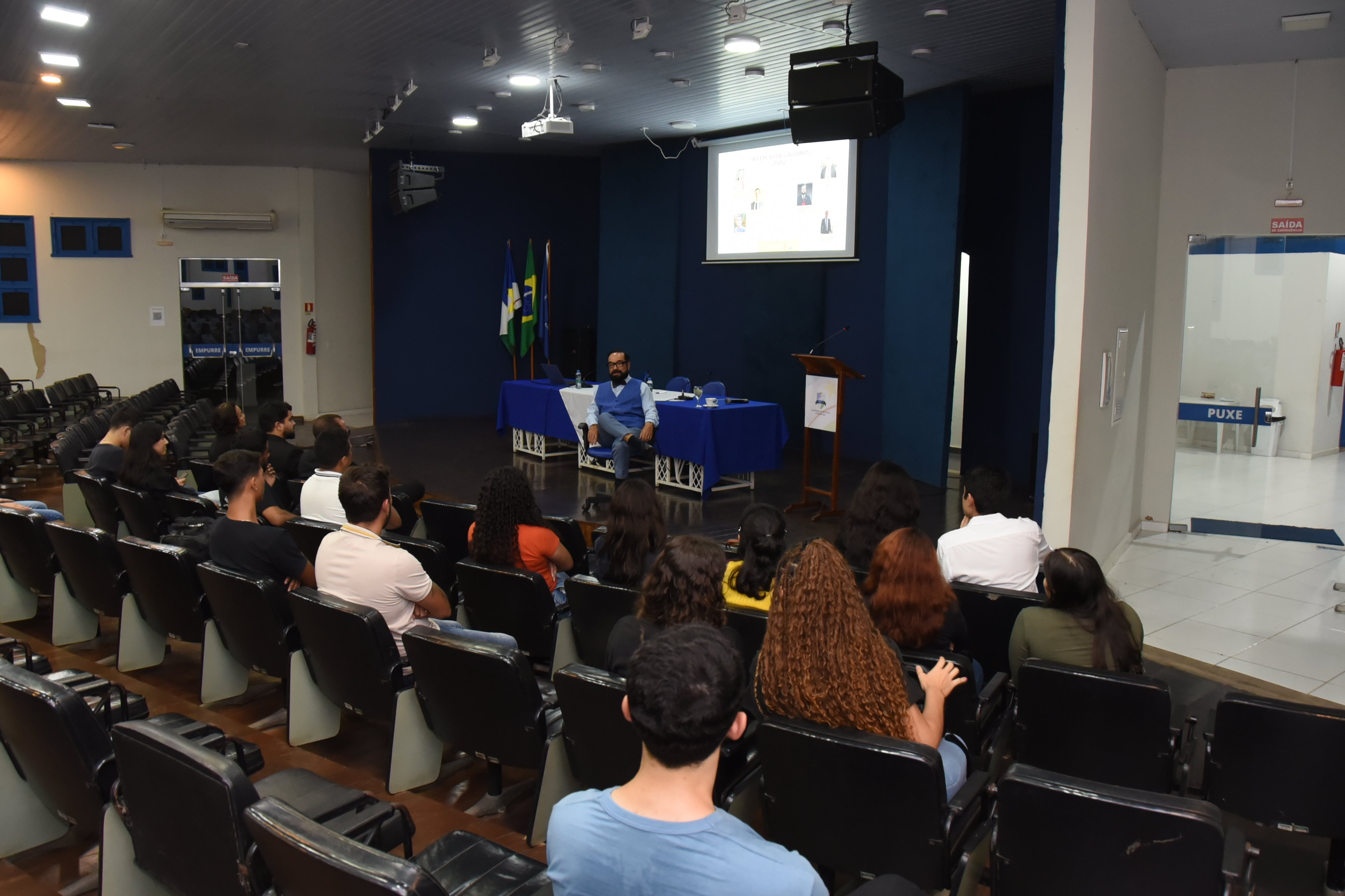 A imagem mostra um auditório com cadeiras pretas ocupadas por jovens atentos a uma palestra. No centro o presidente do TJRR, desembargador Leonardo Cupello, discrusando. O ambiente é fechado, com teto rebaixado, iluminação de LED e paredes nas cores azul e branco. A imagem mostra um auditório com cadeiras pretas ocupadas por jovens atentos a uma palestra. No centro o presidente do TJRR, desembargador Leonardo Cupello, discrusando. O ambiente é fechado, com teto rebaixado, iluminação de LED e paredes nas cores azul e branco.