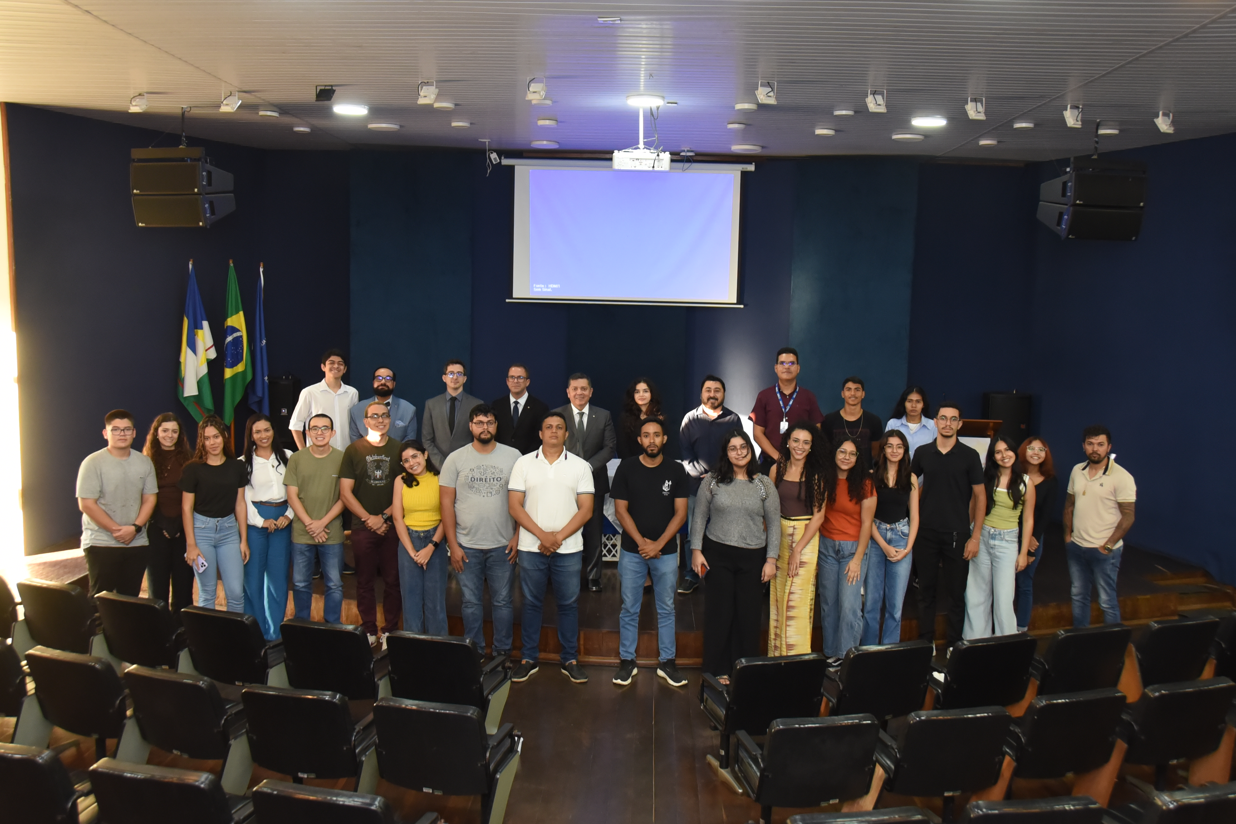A imagem colorida mostra um grupo de 28 reunidas em um auditório para uma foto oficial. Eles estão organizados em duas fileiras em frente ao palco, sorrindo para a câmera. A imagem colorida mostra um grupo de 28 reunidas em um auditório para uma foto oficial. Eles estão organizados em duas fileiras em frente ao palco, sorrindo para a câmera.