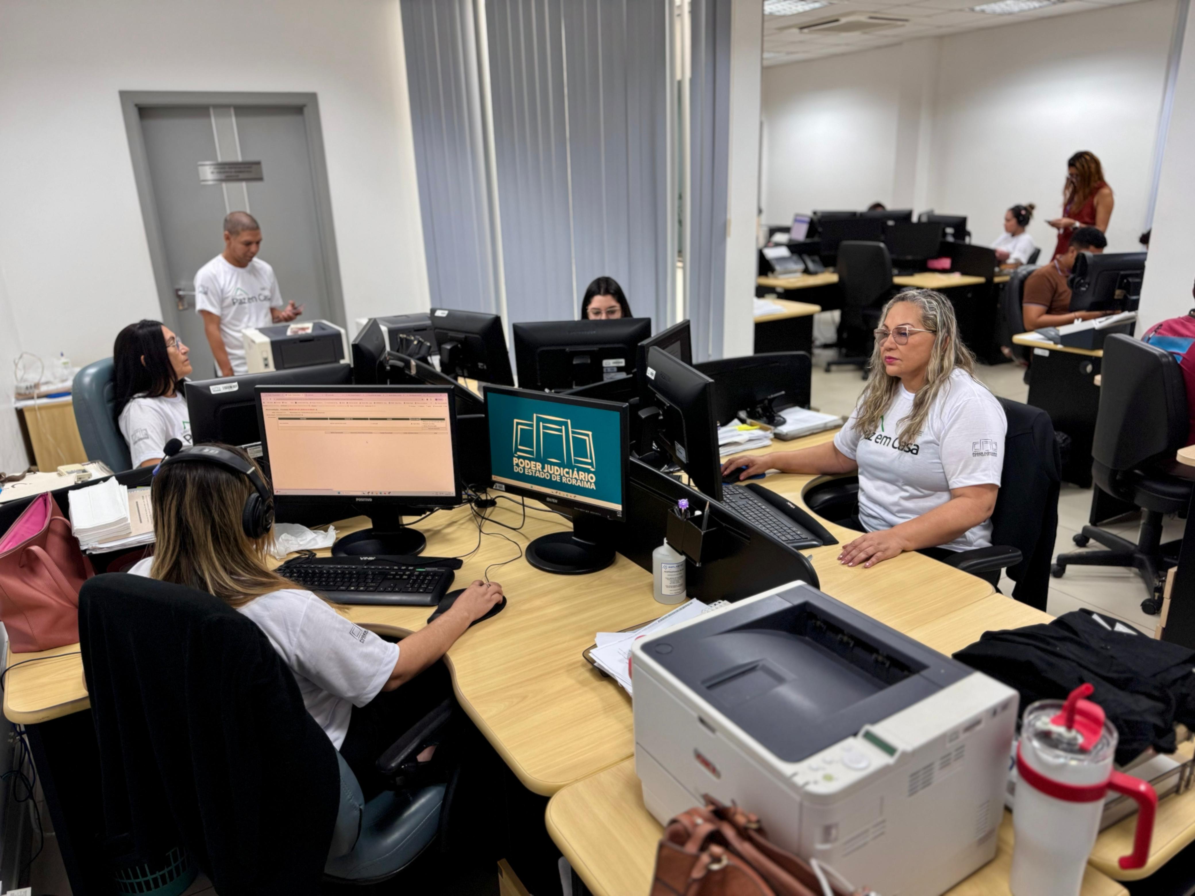 imagem mostra um ambiente de trabalho dando enfoque em cinco pessoas, a maioria mulheres, em suas estações de trabalho. Todos estão vestindo blusas brancas escrito "Paz em casa" imagem mostra um ambiente de trabalho dando enfoque em cinco pessoas, a maioria mulheres, em suas estações de trabalho. Todos estão vestindo blusas brancas escrito "Paz em casa"