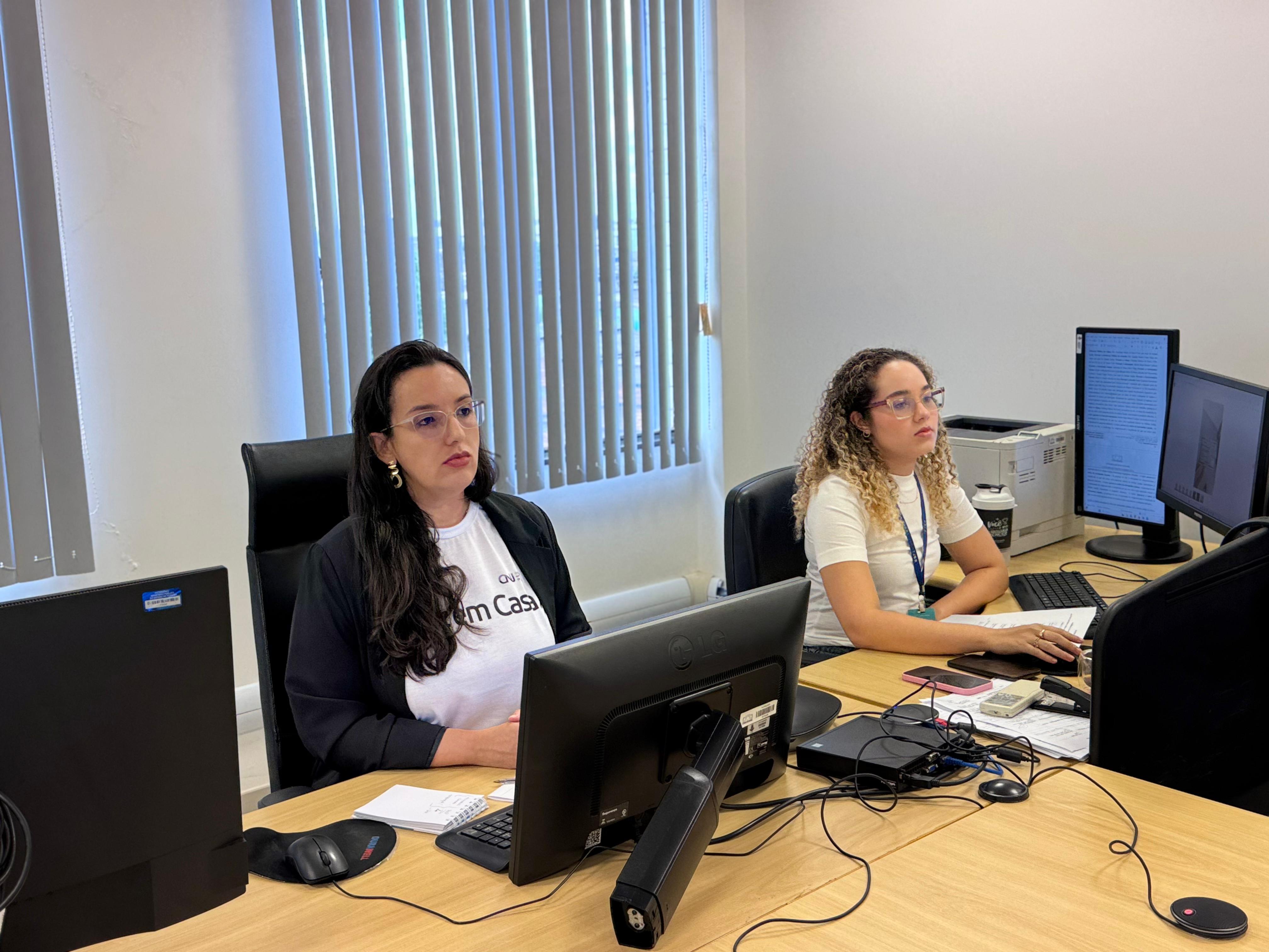 imagem mostra um ambiente de trabalho dando enfoque em duas pessoas, duas mulheres que estão em suas estação de trabalho. Uma está vestindo blusas brancas escrito "Paz em casa". imagem mostra um ambiente de trabalho dando enfoque em duas pessoas, duas mulheres que estão em suas estação de trabalho. Uma está vestindo blusas brancas escrito "Paz em casa".
