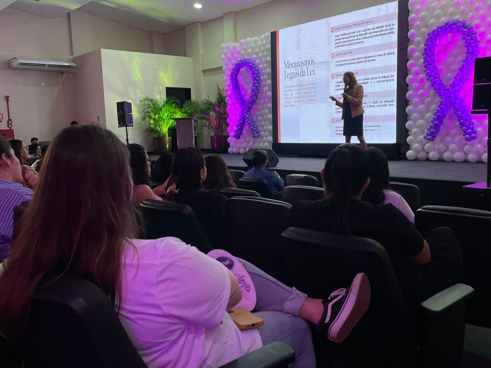 Fotografia mostra a chefe do setor de enfrentamento palestrando em frente a um telão, que exibe o slide intitulado “Mecanismos Legais da Lei”. Ao fundo, o palco está decorado com dois grandes laços lilás formados por balões brancos e roxos, símbolo da campanha de conscientização pelo fim da violência contra a mulher. Na plateia, pessoas sentadas acompanham a apresentação, algumas usando camisetas claras e outras de roupas escuras. Fotografia mostra a chefe do setor de enfrentamento palestrando em frente a um telão, que exibe o slide intitulado “Mecanismos Legais da Lei”. Ao fundo, o palco está decorado com dois grandes laços lilás formados por balões brancos e roxos, símbolo da campanha de conscientização pelo fim da violência contra a mulher. Na plateia, pessoas sentadas acompanham a apresentação, algumas usando camisetas claras e outras de roupas escuras.