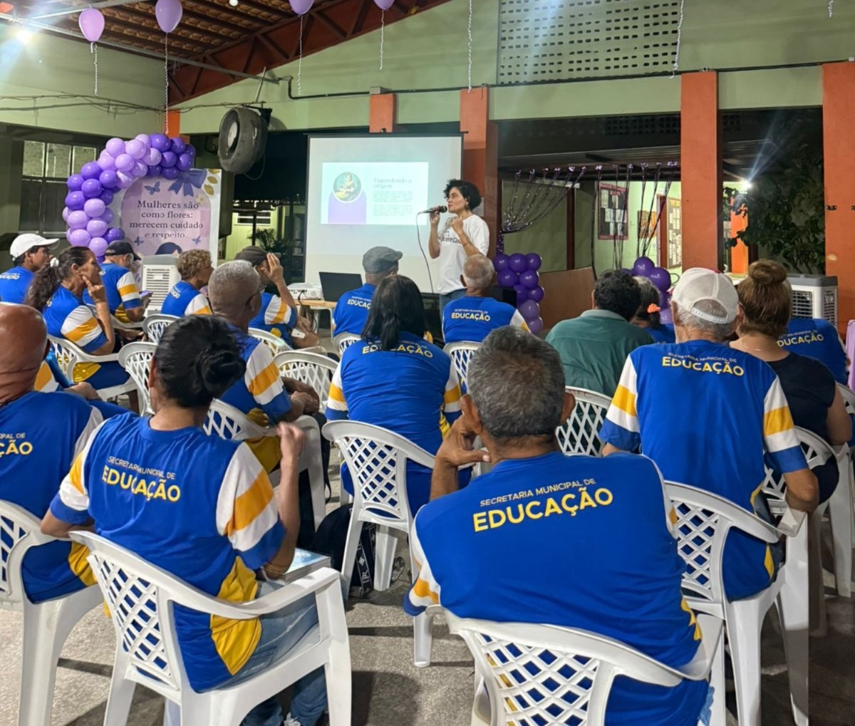 Fotografia mostra uma palestra realizada em um espaço fechado. No centro, uma mulher em pé, usando camiseta branca, fala ao público com microfone em mãos, em frente a uma tela de projeção que exibe um slide. O auditório está decorado com balões lilás e uma faixa onde se lê: “Mulheres são como flores: merecem cuidado e respeito”. As cadeiras brancas estão ocupadas por participantes, a maioria vestindo camisas azuis com detalhes amarelos e brancos, onde está escrito “Secretaria Municipal de Educação”. Fotografia mostra uma palestra realizada em um espaço fechado. No centro, uma mulher em pé, usando camiseta branca, fala ao público com microfone em mãos, em frente a uma tela de projeção que exibe um slide. O auditório está decorado com balões lilás e uma faixa onde se lê: “Mulheres são como flores: merecem cuidado e respeito”. As cadeiras brancas estão ocupadas por participantes, a maioria vestindo camisas azuis com detalhes amarelos e brancos, onde está escrito “Secretaria Municipal de Educação”.