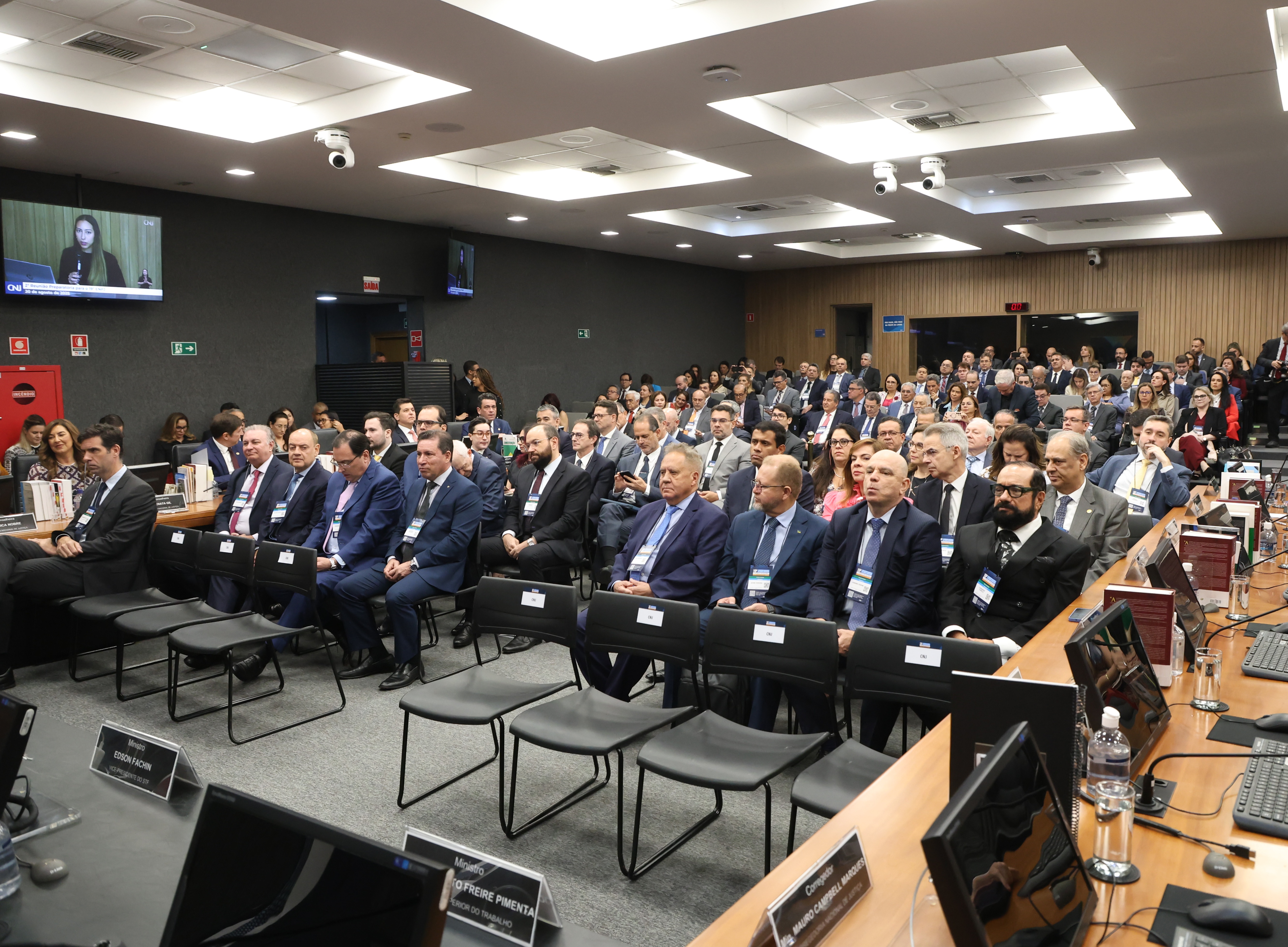Foto colorida mostra um auditório lotado de autoridades assistindo a uma solenidade, entre eles o presidente do TJRR, Desembargador Leonardo Cupello. À frente, algumas cadeiras estão reservadas com identificações nominais. No ambiente, é possível ver monitores de computador sobre mesas, câmeras de segurança no teto e televisores transmitindo a programação de um canal de notícias. Foto colorida mostra um auditório lotado de autoridades assistindo a uma solenidade, entre eles o presidente do TJRR, Desembargador Leonardo Cupello. À frente, algumas cadeiras estão reservadas com identificações nominais. No ambiente, é possível ver monitores de computador sobre mesas, câmeras de segurança no teto e televisores transmitindo a programação de um canal de notícias.